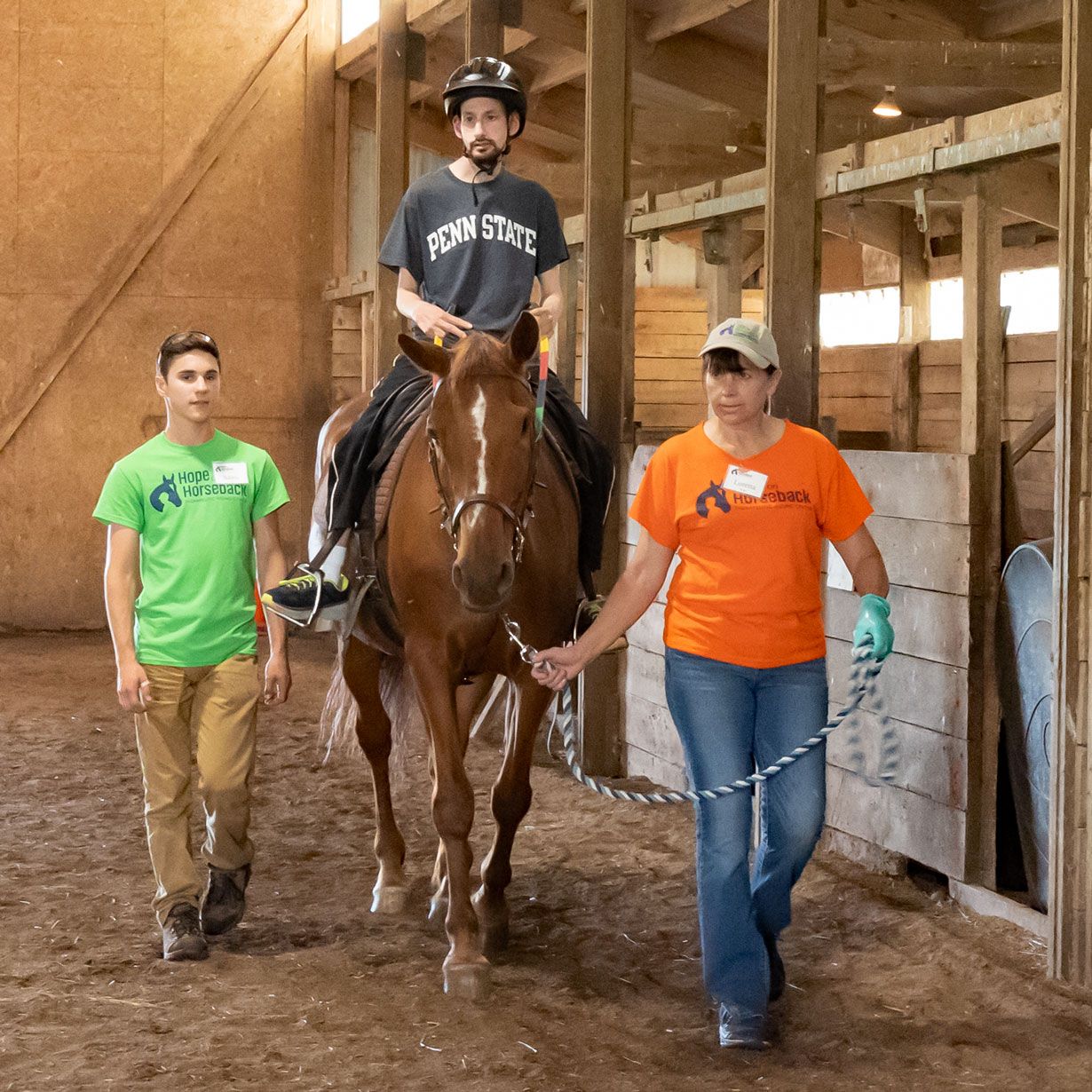 Person on brown horse led by two handlers in a barn aisle, one in green shirt and one in orange shirt