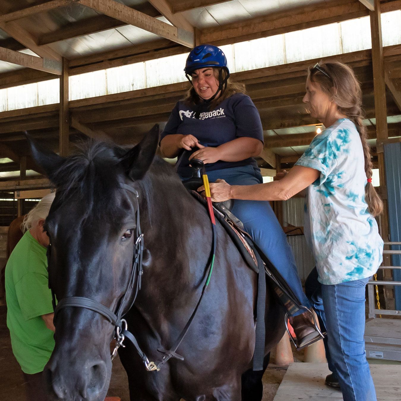 Rider on a black horse indoors while a helper adjusts the reins beside the saddle