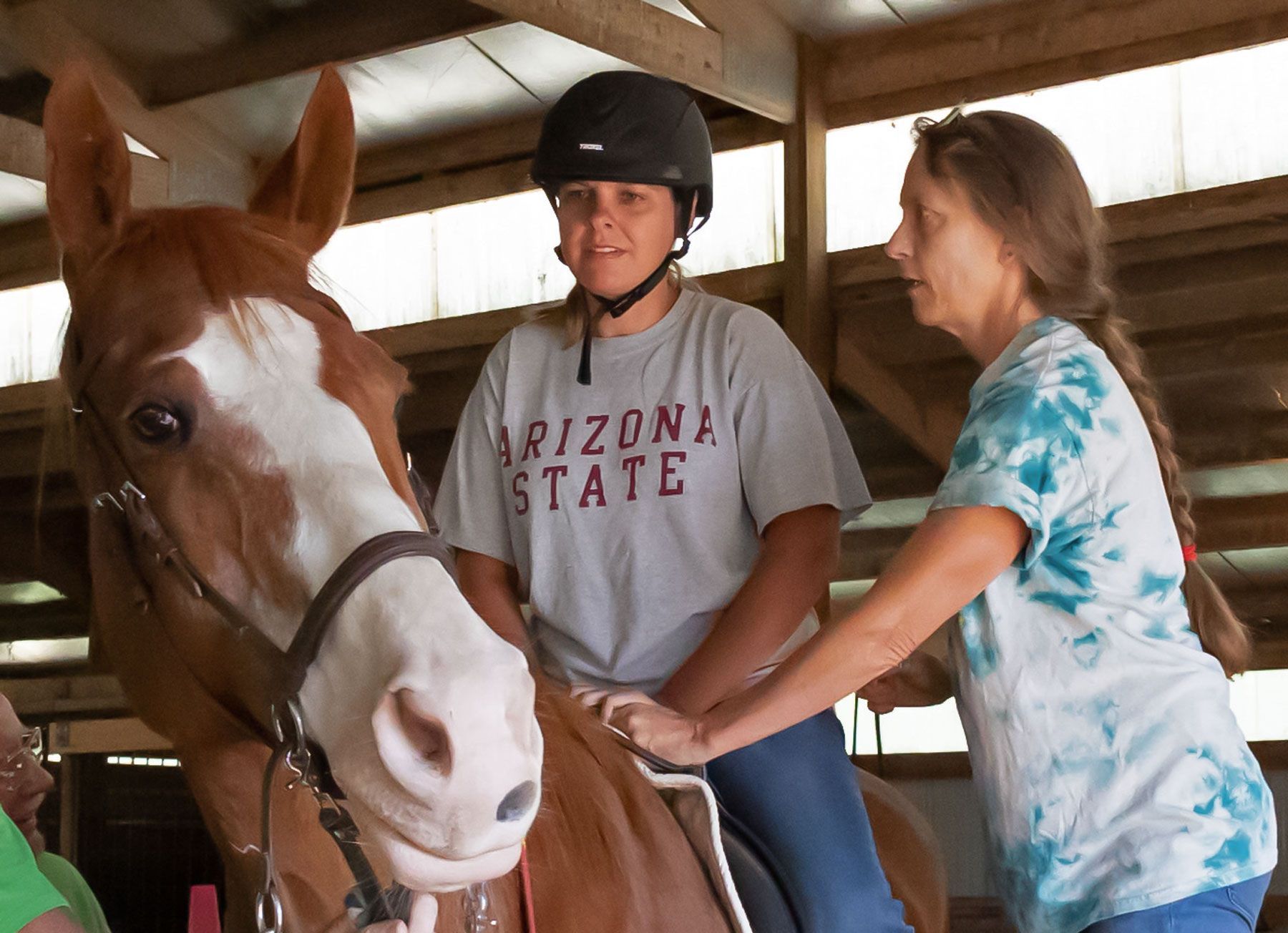 Person in helmet riding a brown horse while another woman helps them in a stable