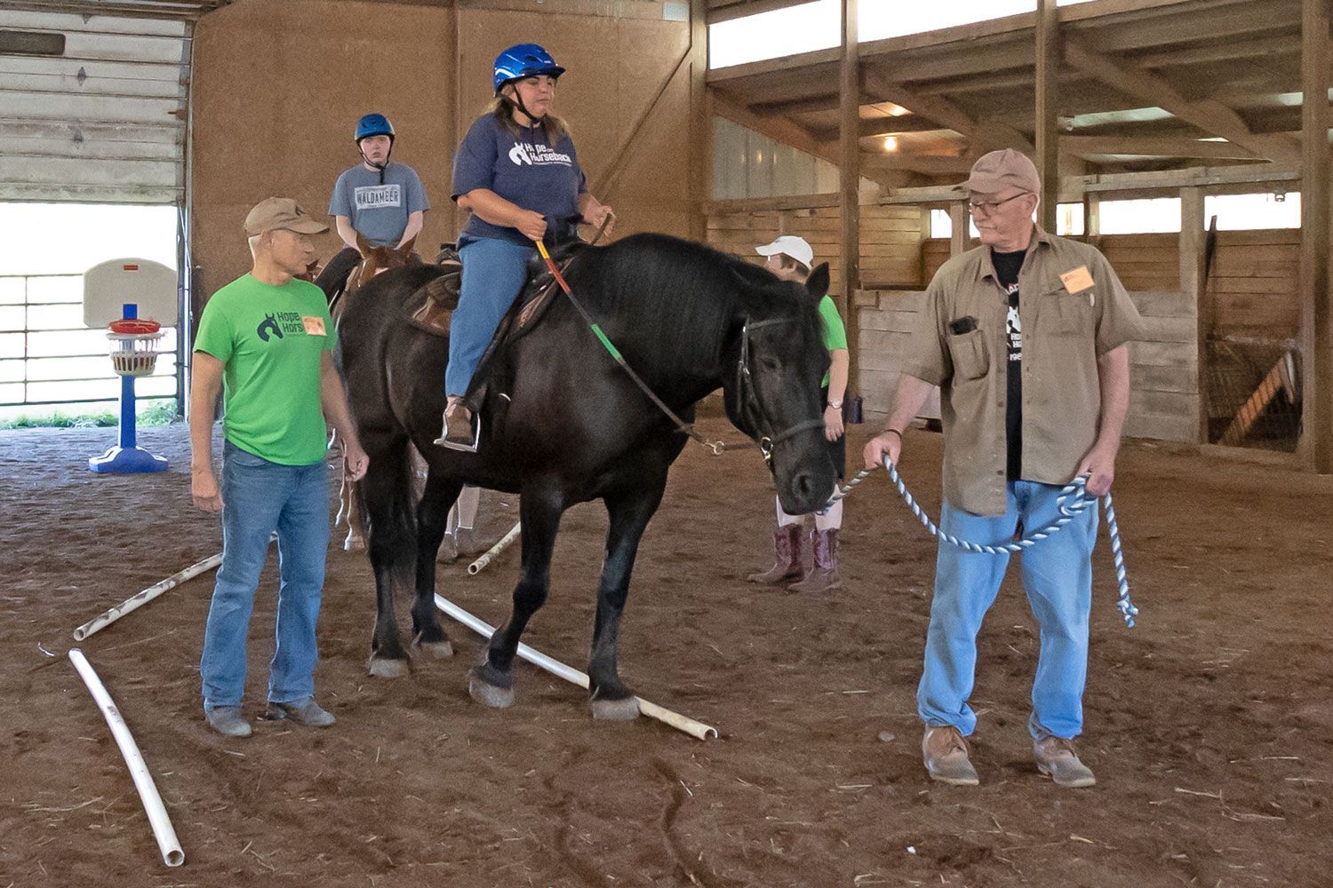 Person on black horse in indoor riding arena, with two handlers nearby and a child riding behind.