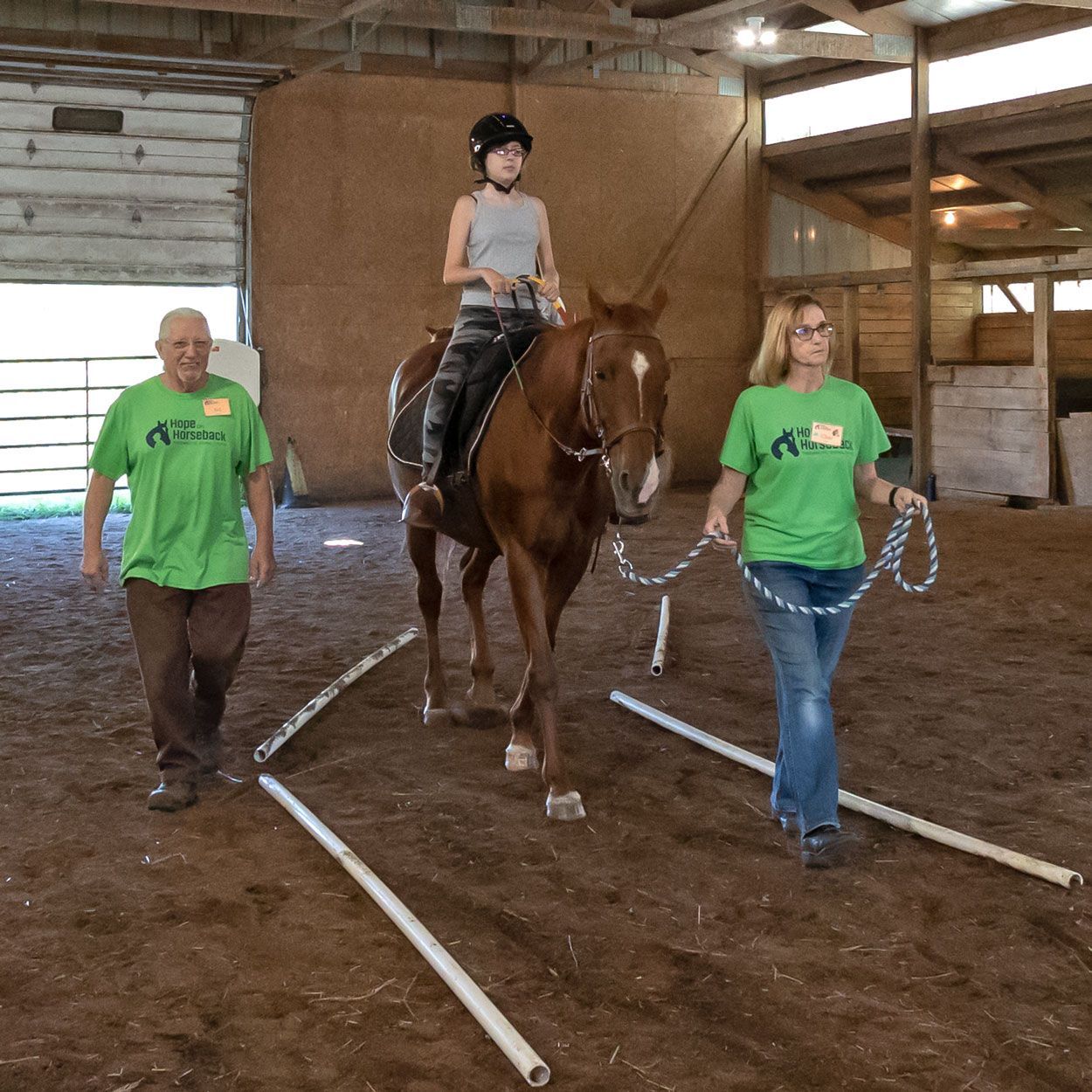 Indoor horse-riding lesson with a rider on a brown horse between two handlers in a barn.