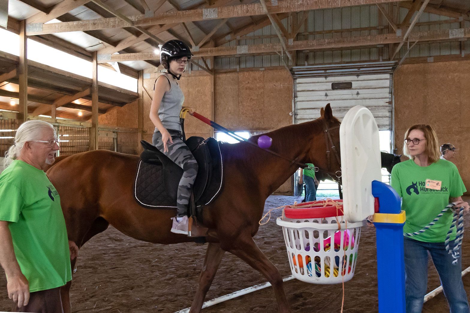 Girl riding brown horse in barn while two adults stand nearby and watch.