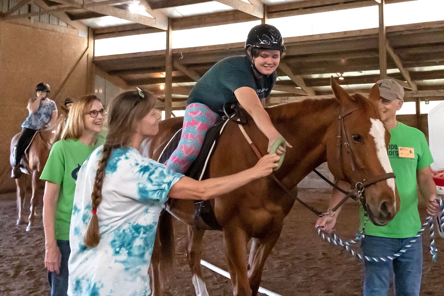 Person riding a brown horse indoors while others in green shirts guide and support her