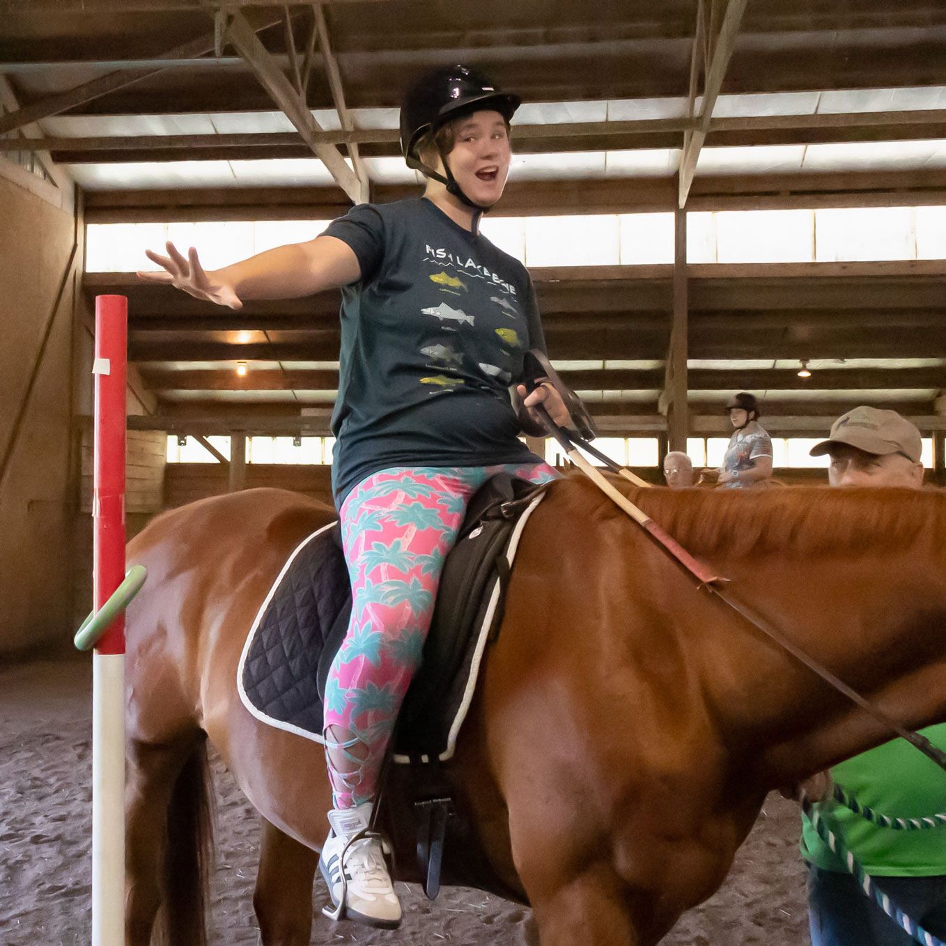 Person riding a brown horse indoors, smiling with one arm outstretched in a riding arena.