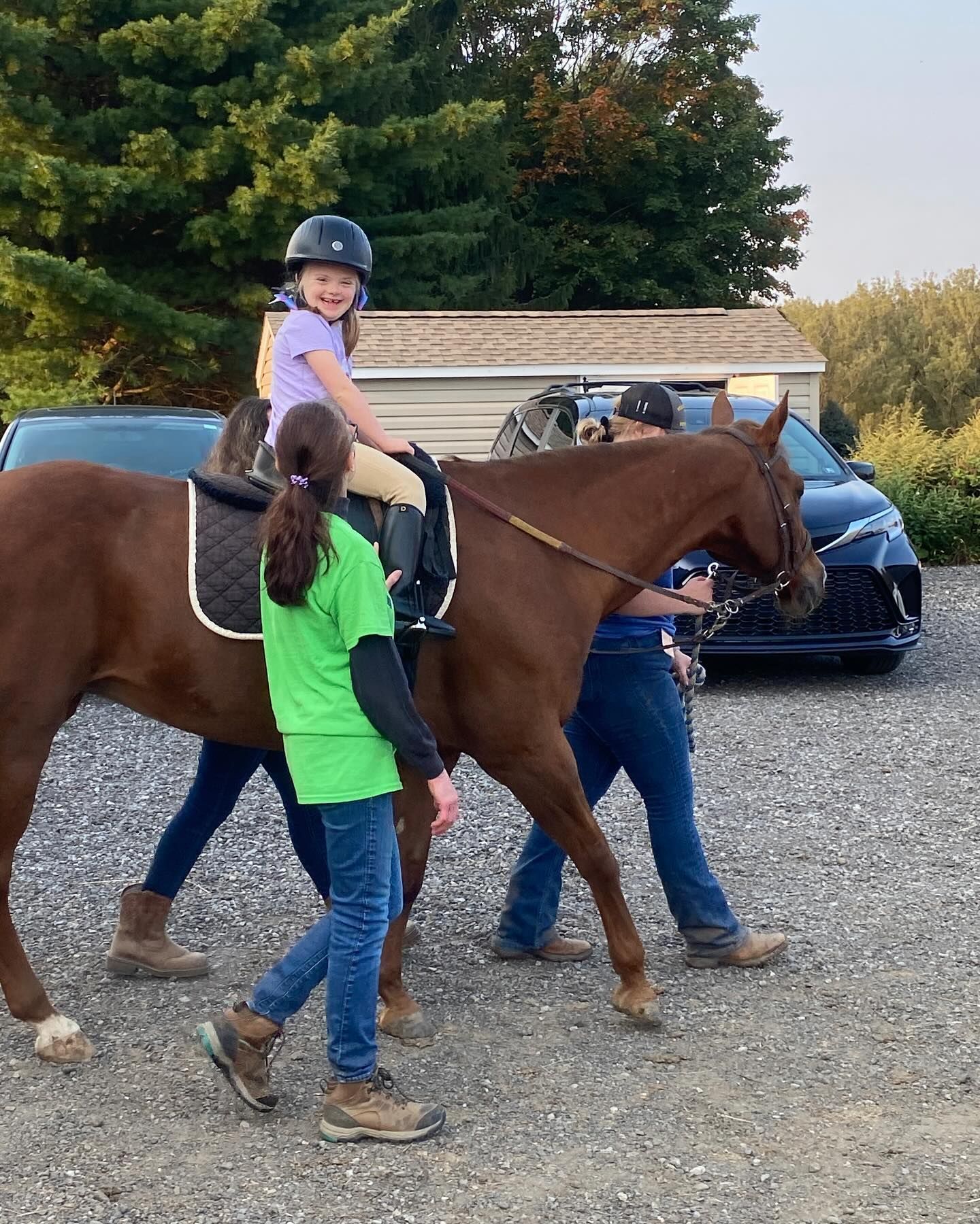 Person leading a brown horse while a child rides it, outdoors on a gravel path.