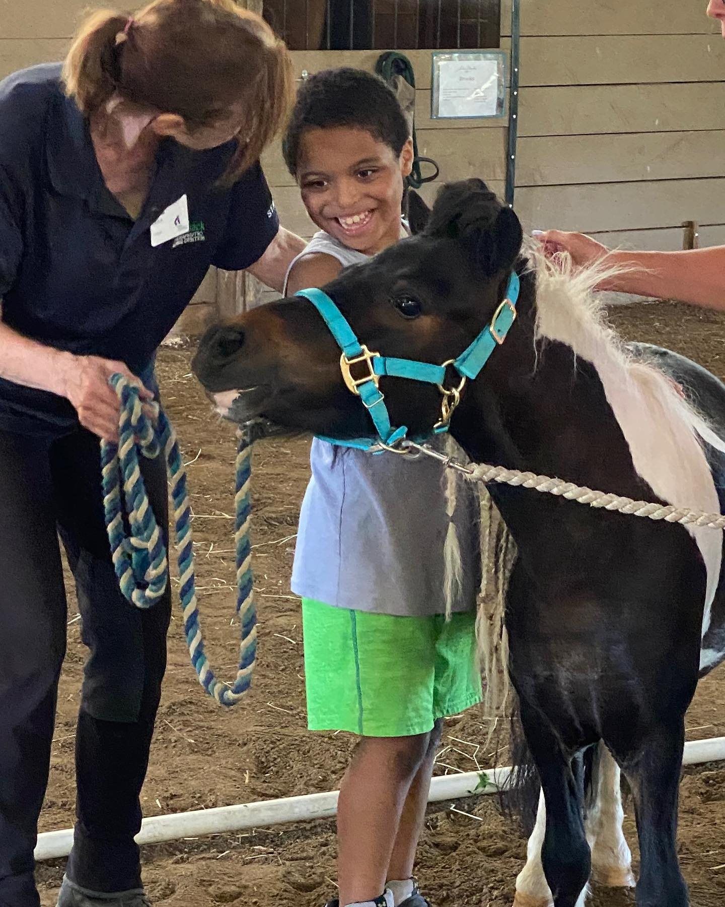 Child smiling beside a small black horse while an adult adjusts the rope halter in a barn.