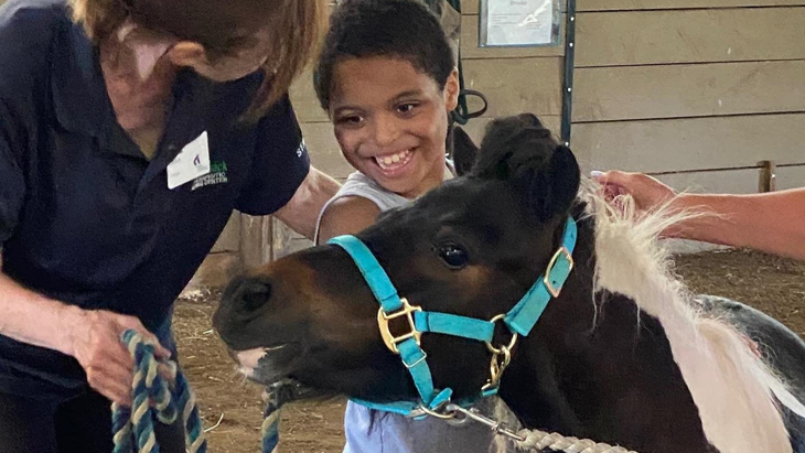 Person smiling beside a small black-and-white pony wearing a turquoise halter.
