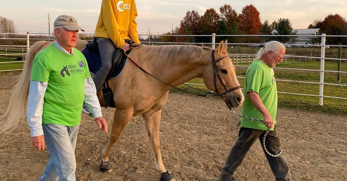 Two people in green shirts lead a tan horse with a rider in a yellow shirt in a fenced paddock at sunset