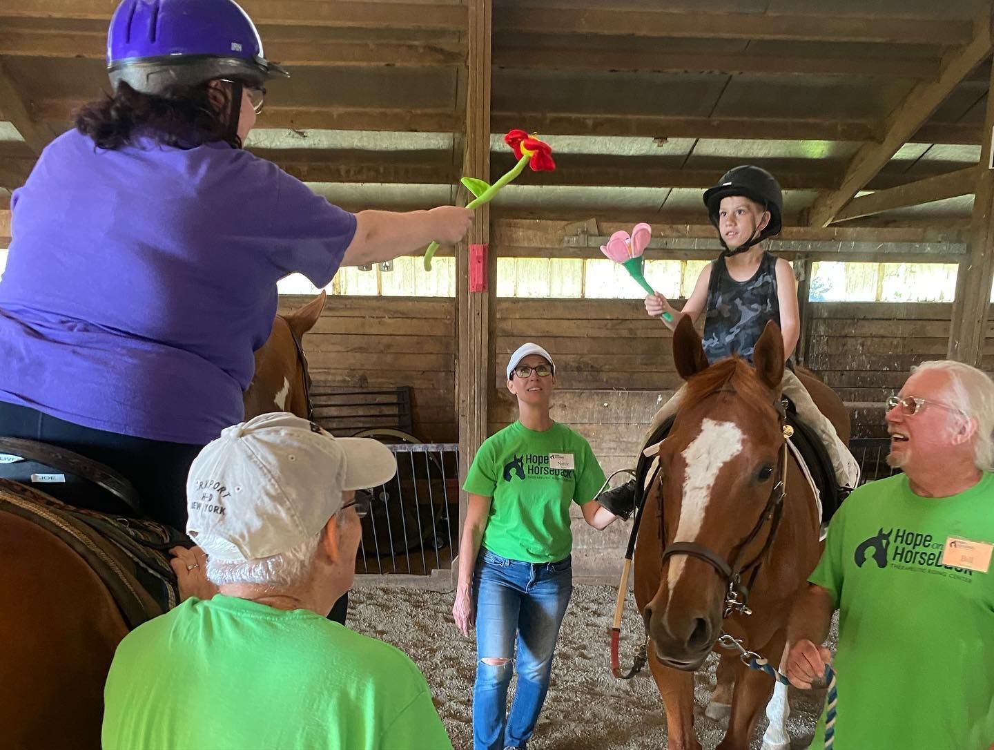 Person on horseback reaches toward a red flower during an indoor stable activity while others watch.