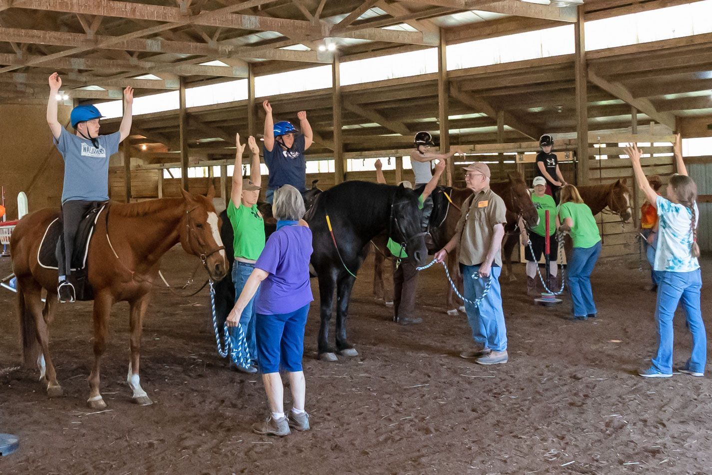 People raising hands around horses in a barn arena, with one person mounted on a brown horse.
