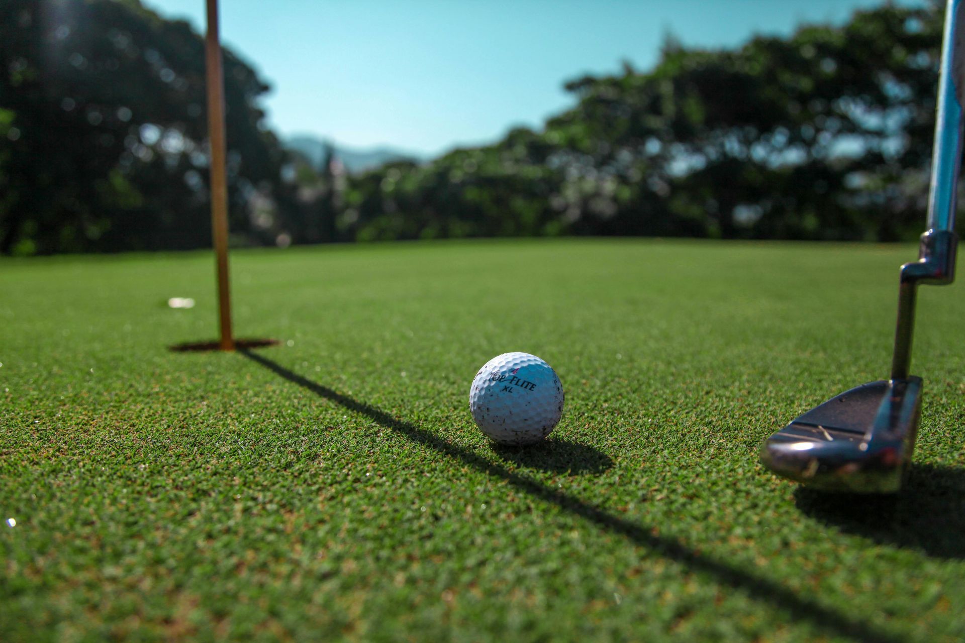Golf ball on green putting course beside flag and putter in sunlight