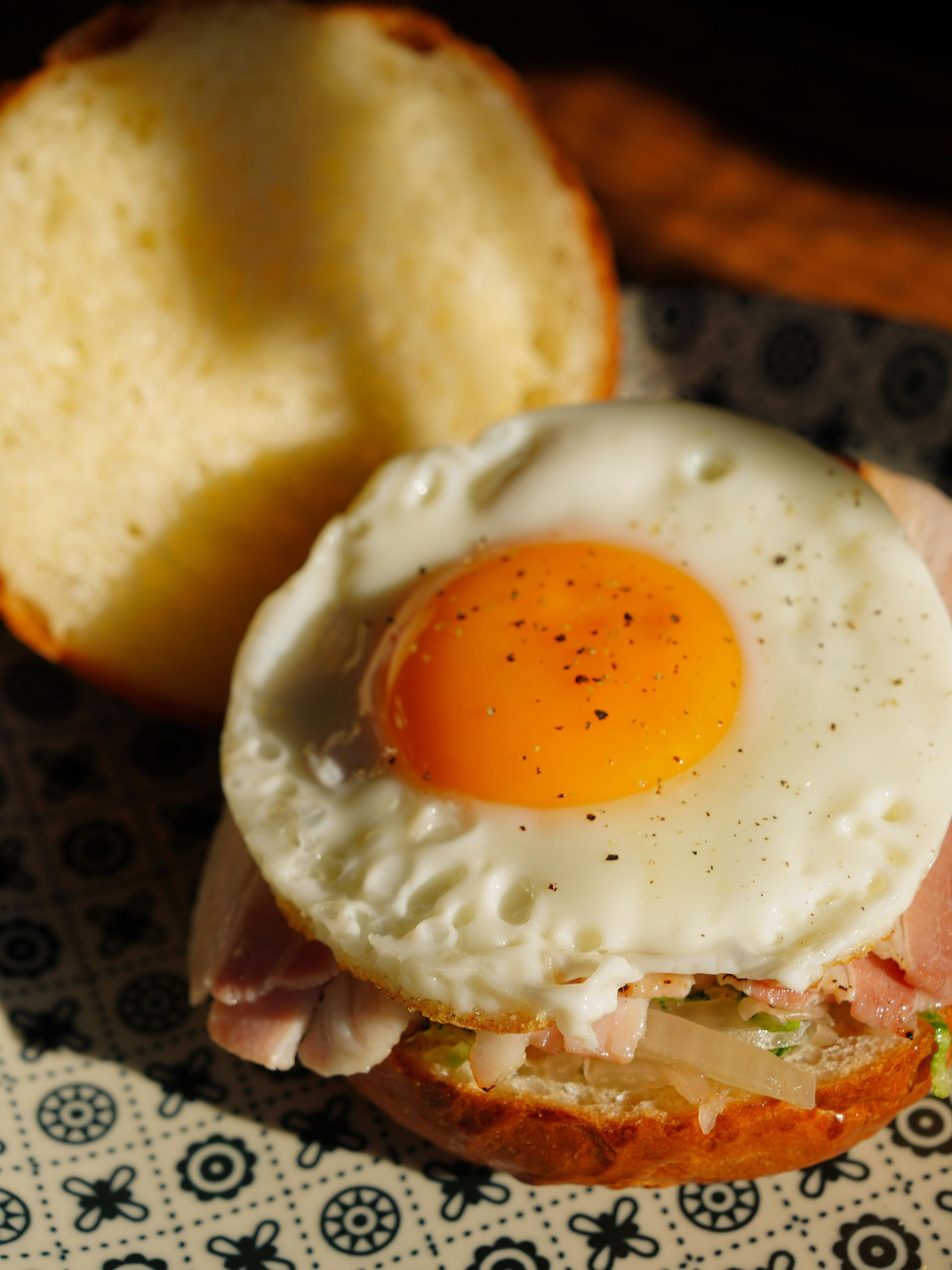 Fried egg on a breakfast sandwich with lettuce and bacon on a bun, with another bun half beside it.