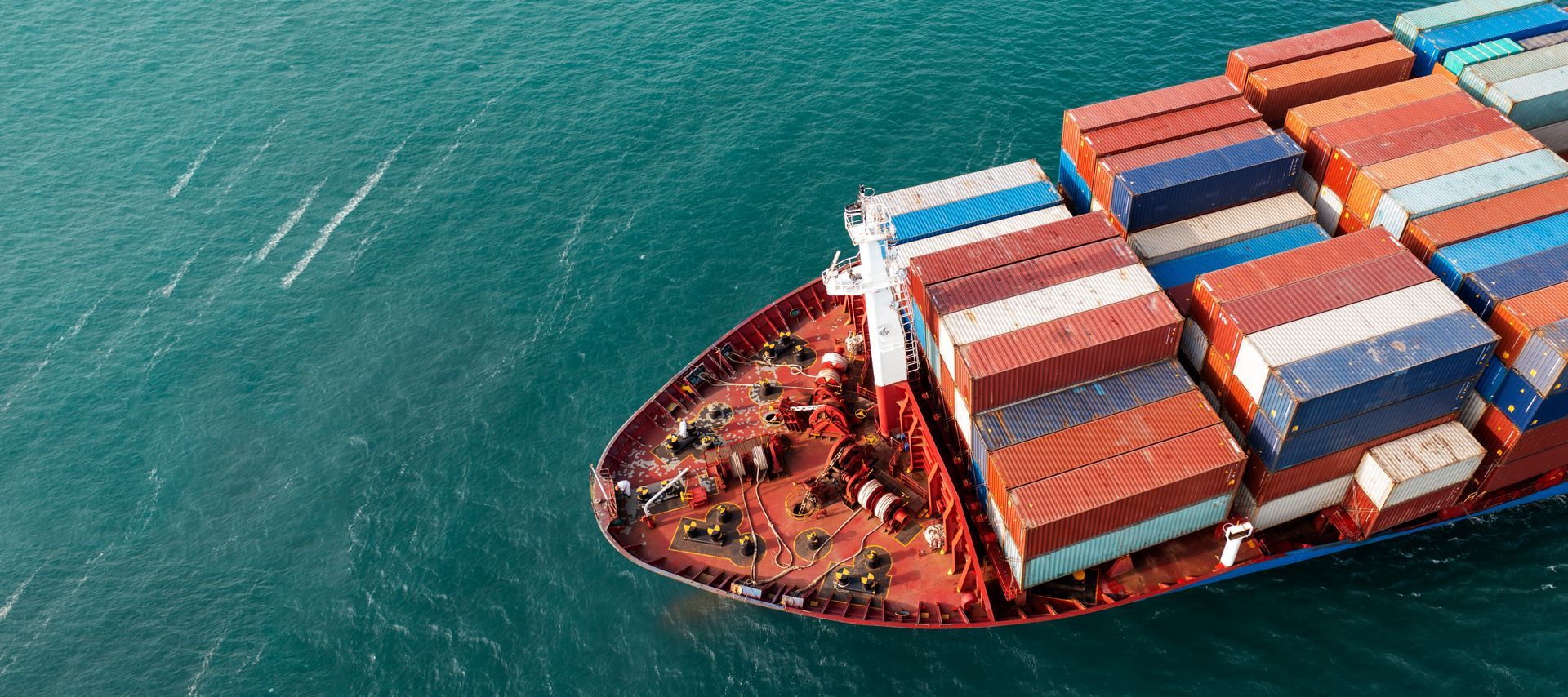 An aerial view of a large cargo ship traveling through open water, carrying rows of colorful shipping containers.