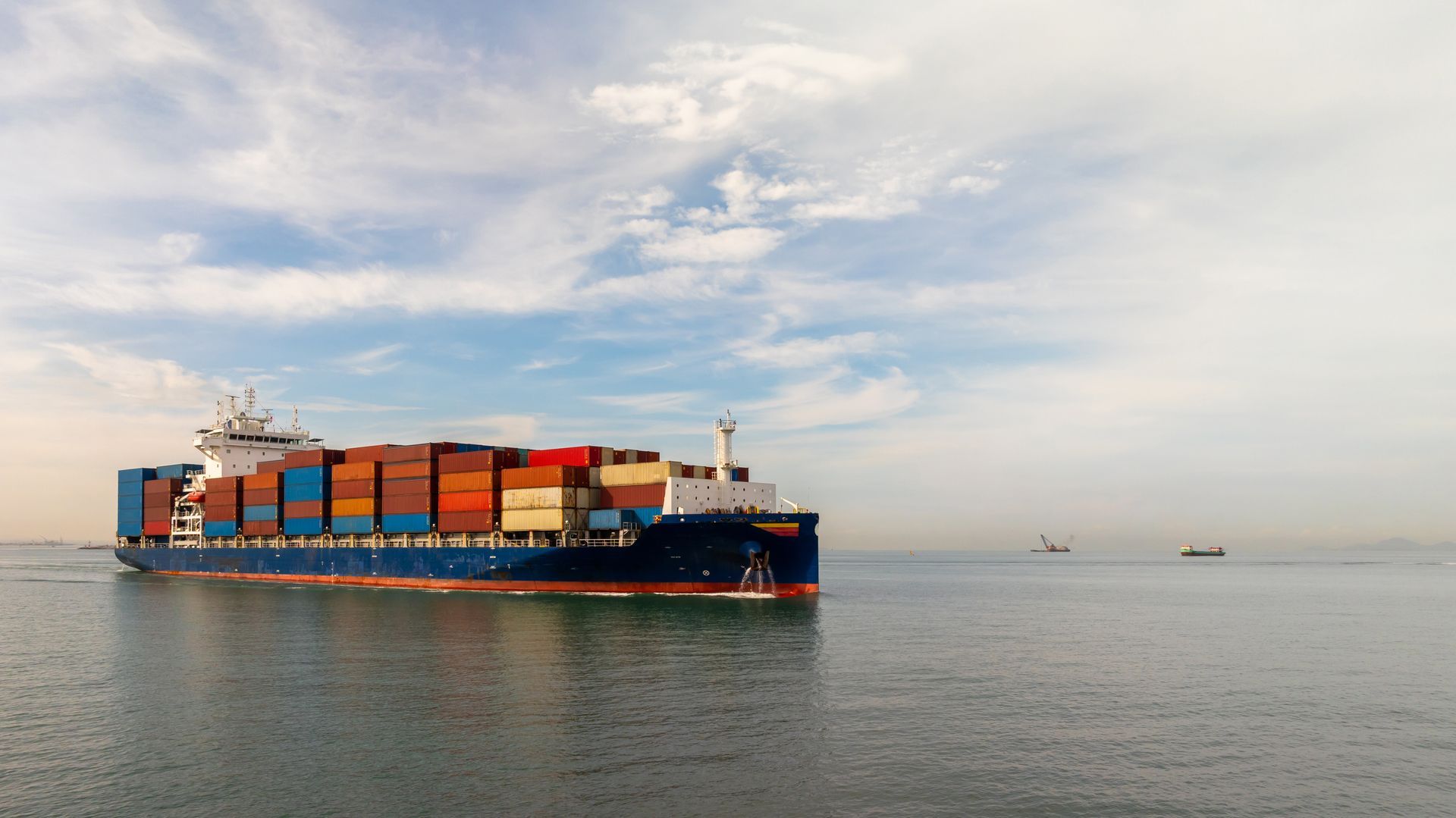 A large container ship loaded with colorful shipping crates moves across a calm, open ocean under a bright, cloudy sky.