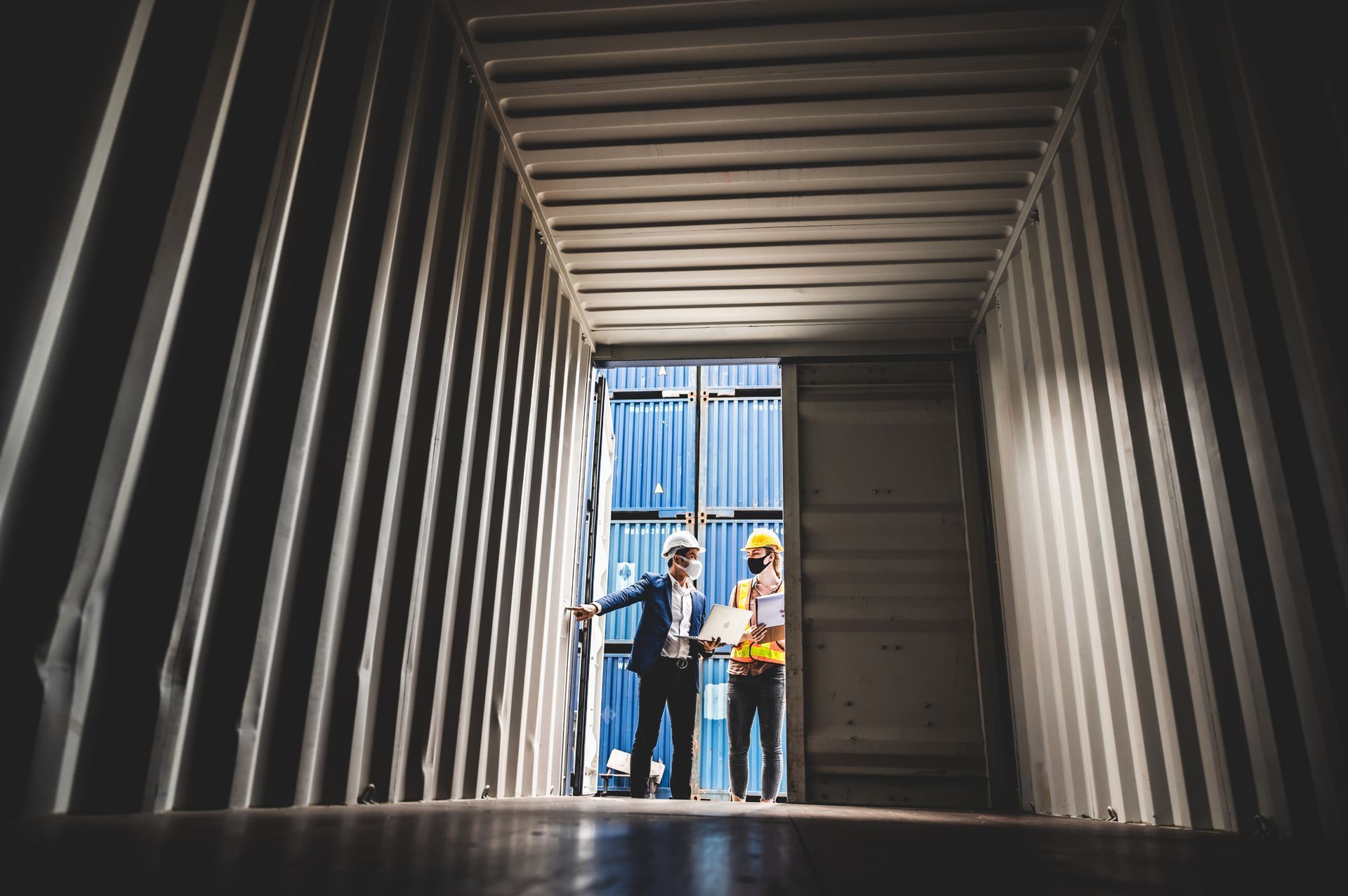 Two professionals in hard hats and masks confer inside a shipping container, backlit by bright daylight.