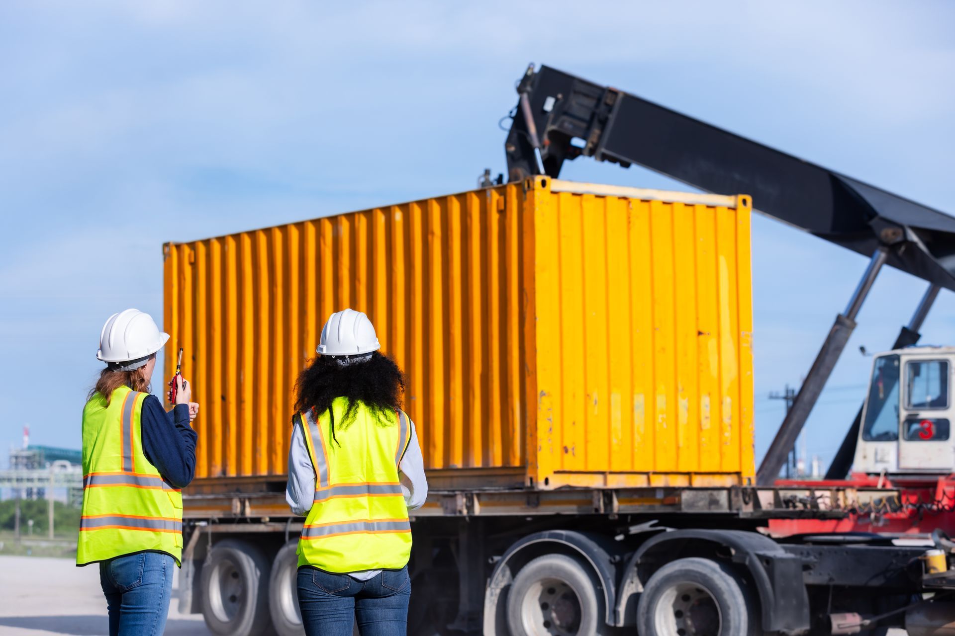 Two workers in hard hats and safety vests stand near a truck loading a yellow shipping container at an industrial site.