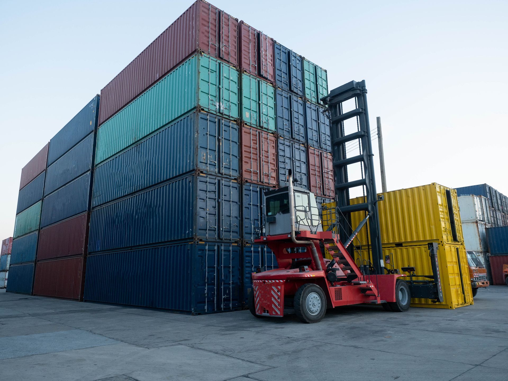 A red reach stacker maneuvering a yellow shipping container next to a large, multi-colored stack of containers.
