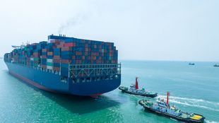A large container ship sailing on the sea, guided by two tugboats on its starboard side.