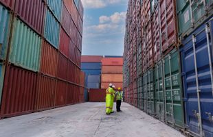 Two workers in safety gear walk between two towering, colorful stacks of shipping containers in a yard.