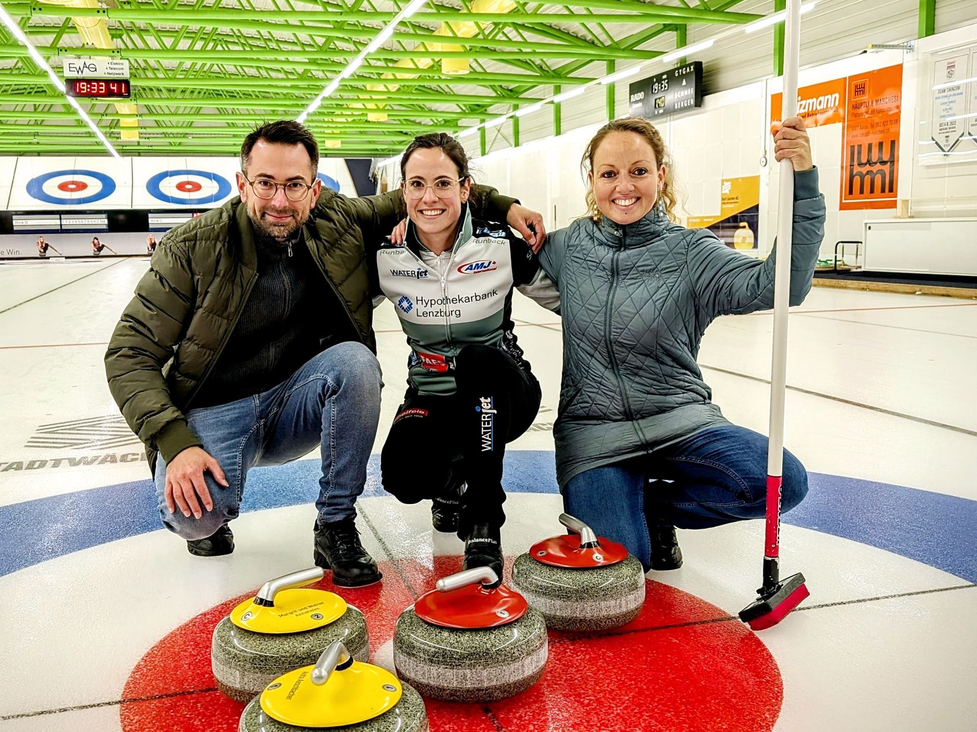 Drei Personen die auf einem Curlingfeld knien und für das Foto posieren. Eine davon hält den Besen.