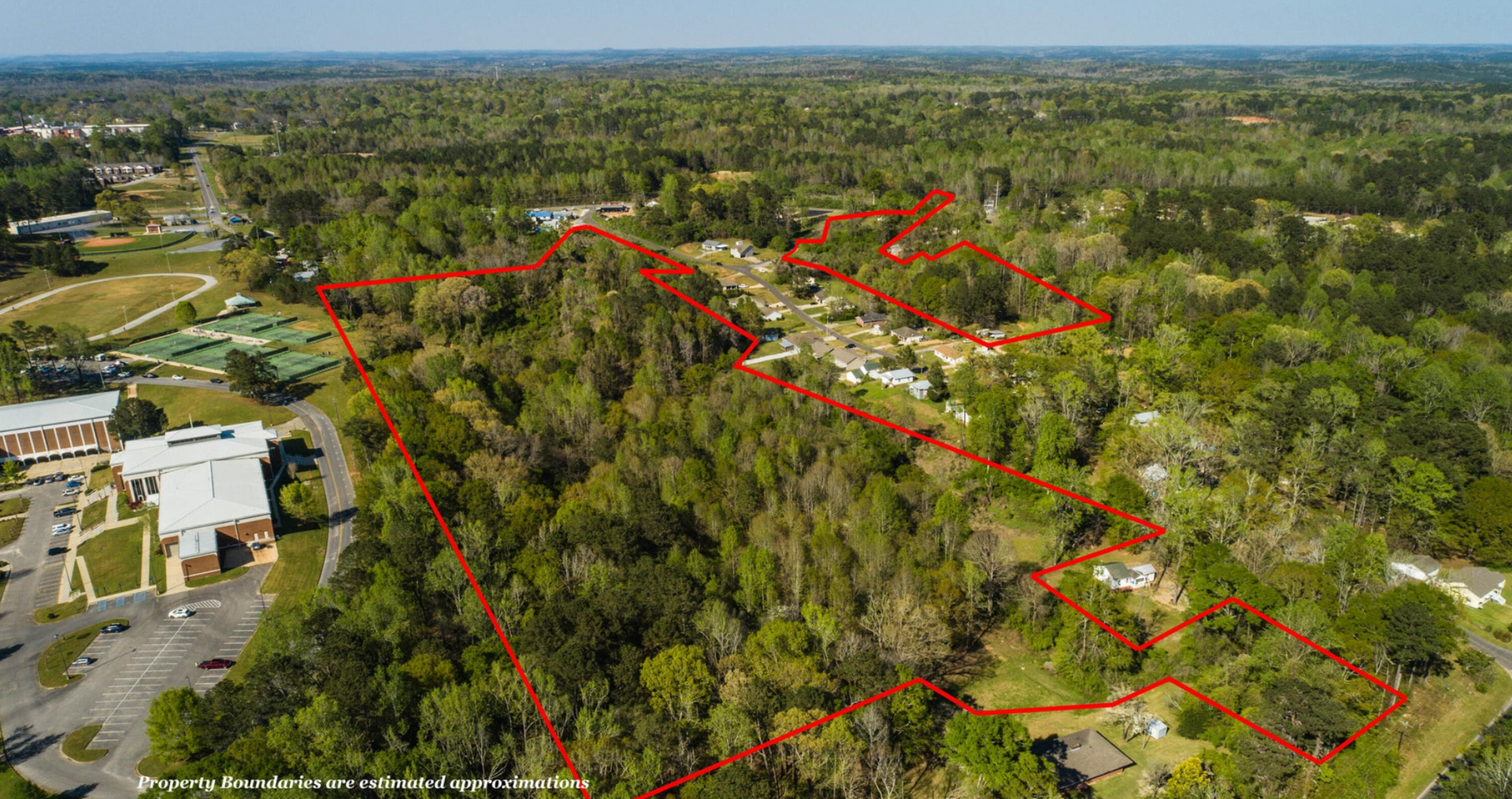 Aerial view of a wooded lot marked with a red outline, situated next to buildings and a parking area.