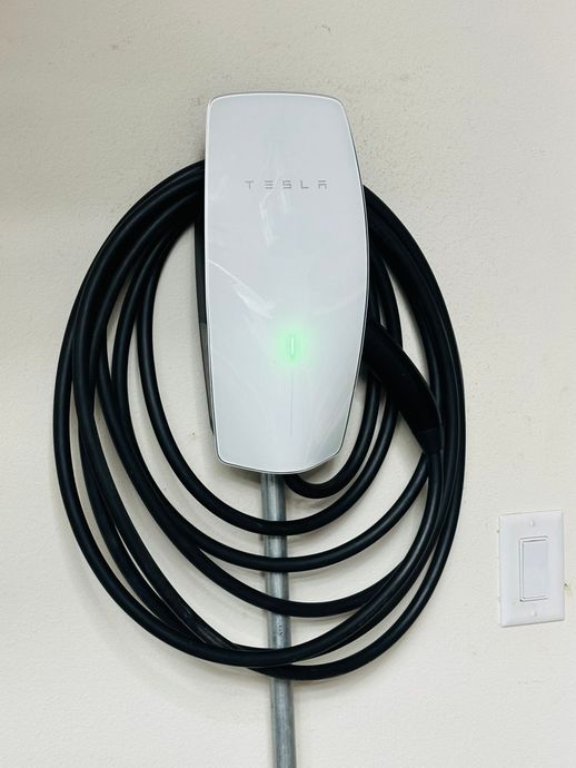 White EV wall charger with coiled black cable mounted on a wall beside a light switch.