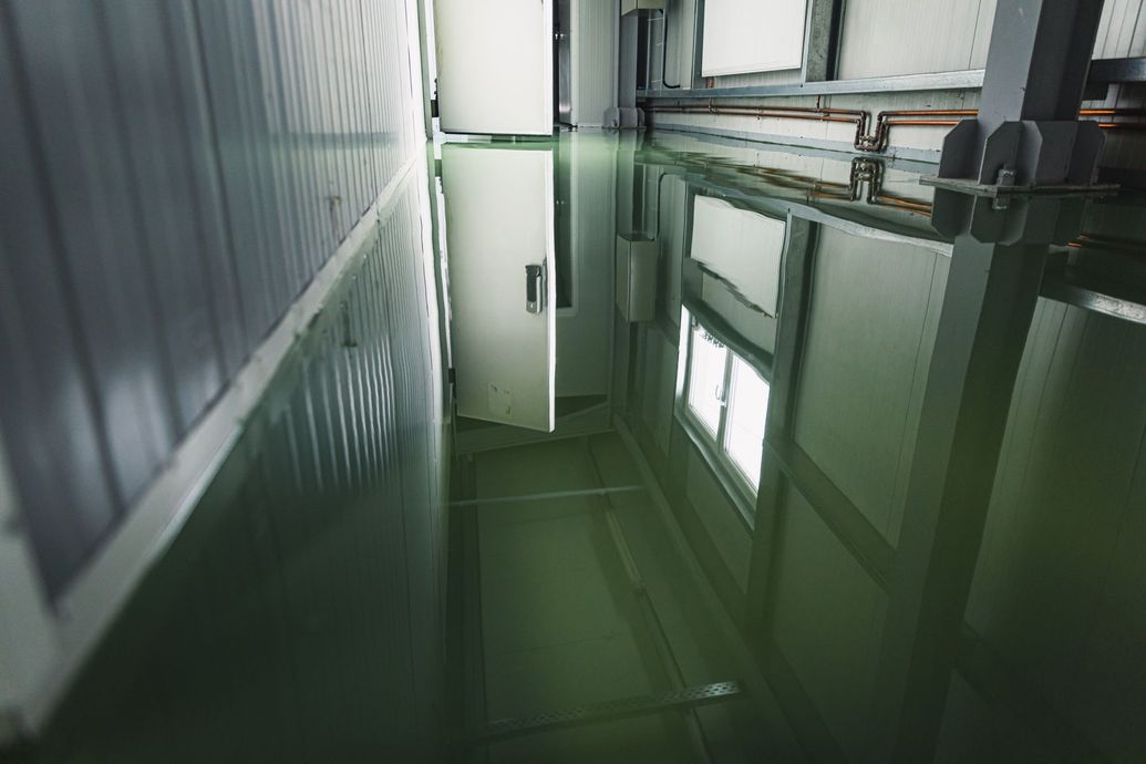 Narrow green-tinted industrial corridor with windows, pipes, and reflective walls.