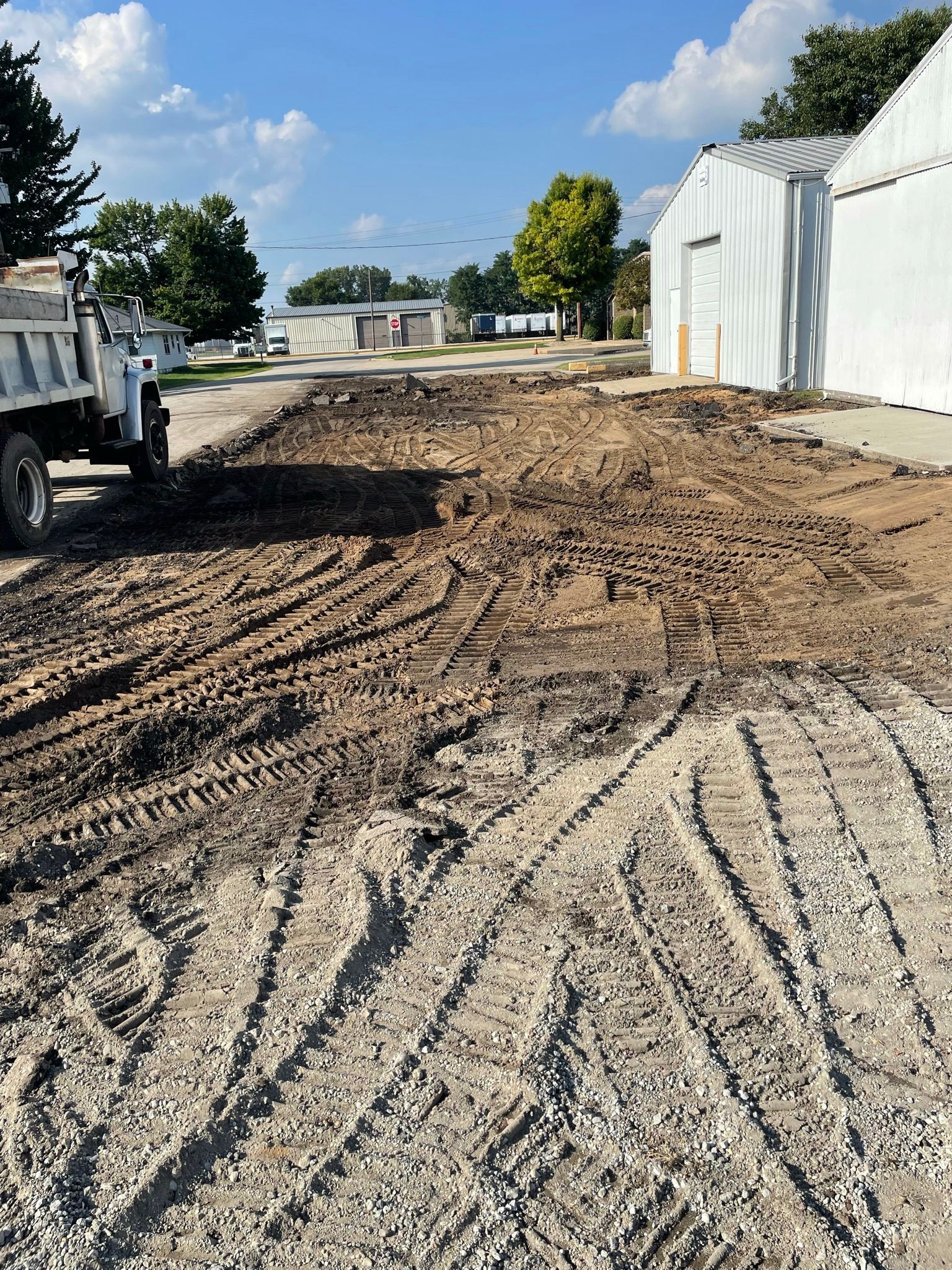 Muddy ground with tire tracks next to a building and a dump truck under a blue sky.