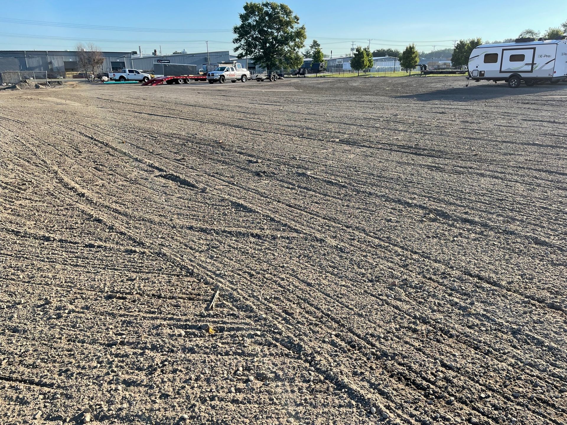 Gravel parking lot with tire tracks. Vehicles and a trailer parked in the background. Clear sky.
