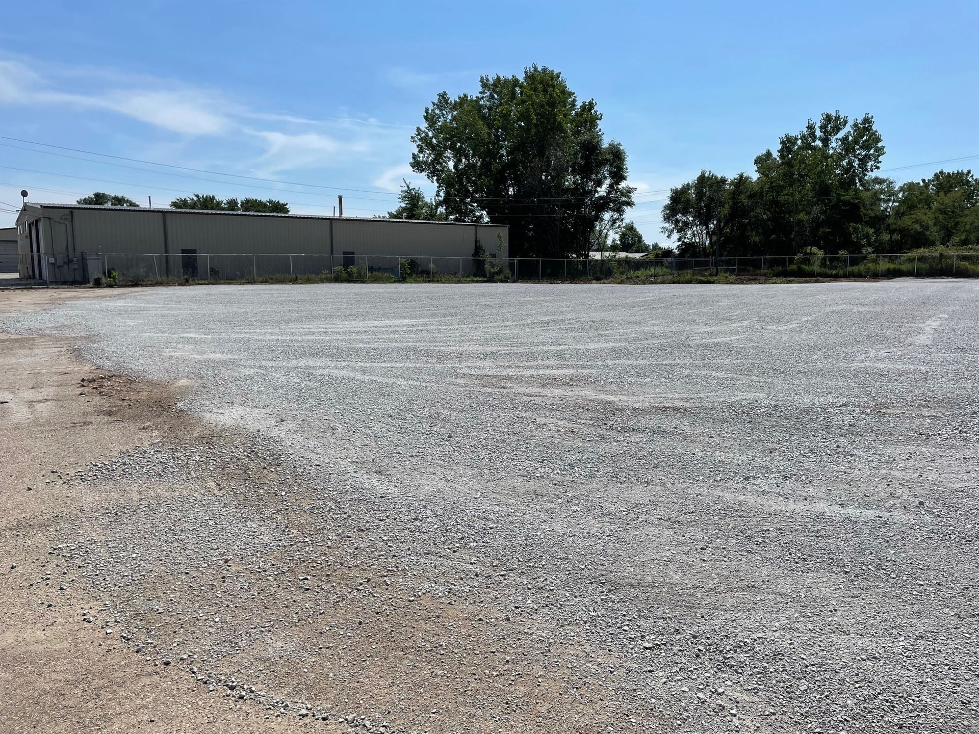 Gravel lot in front of a low gray building, with trees and a clear blue sky.