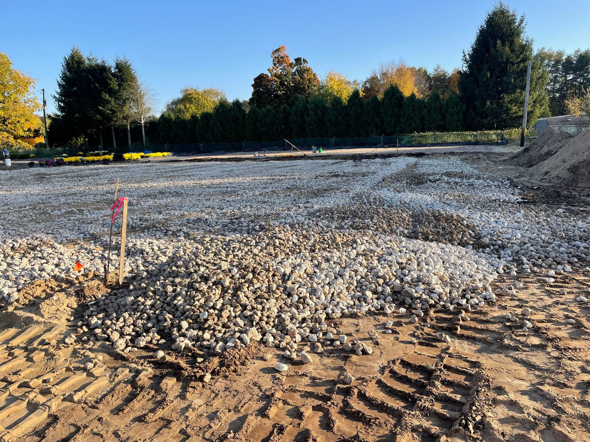 Construction site with gravel covering a cleared area, tire tracks in dirt, trees in background.
