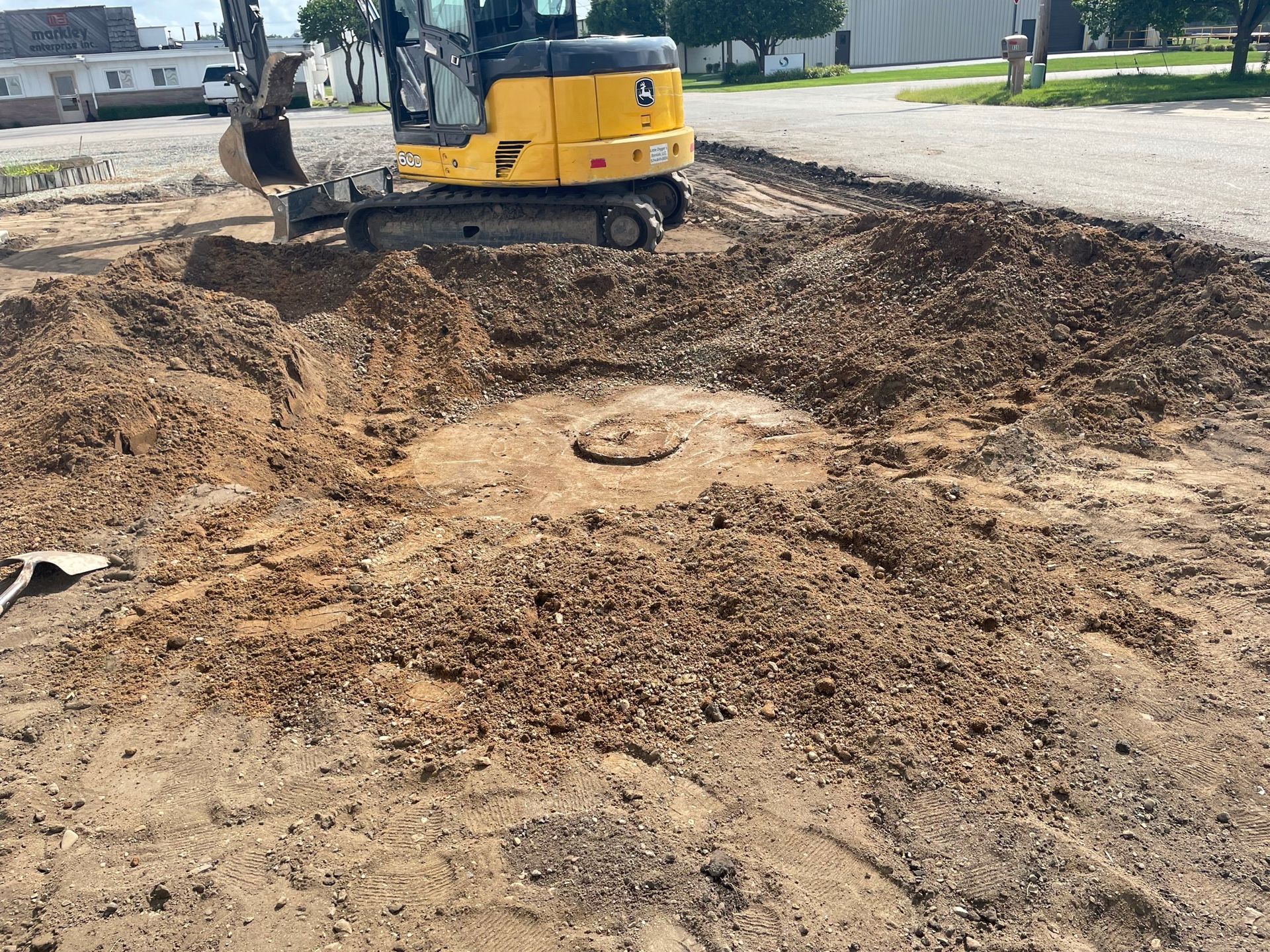 A yellow excavator digs around a buried manhole cover in a dirt lot.
