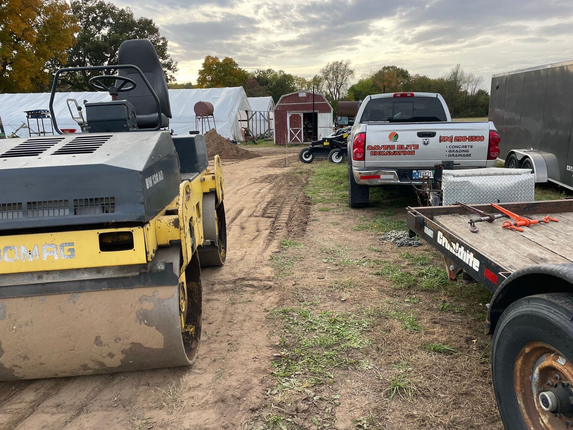 Yellow road roller, truck, trailer, and small buildings in a rural area.