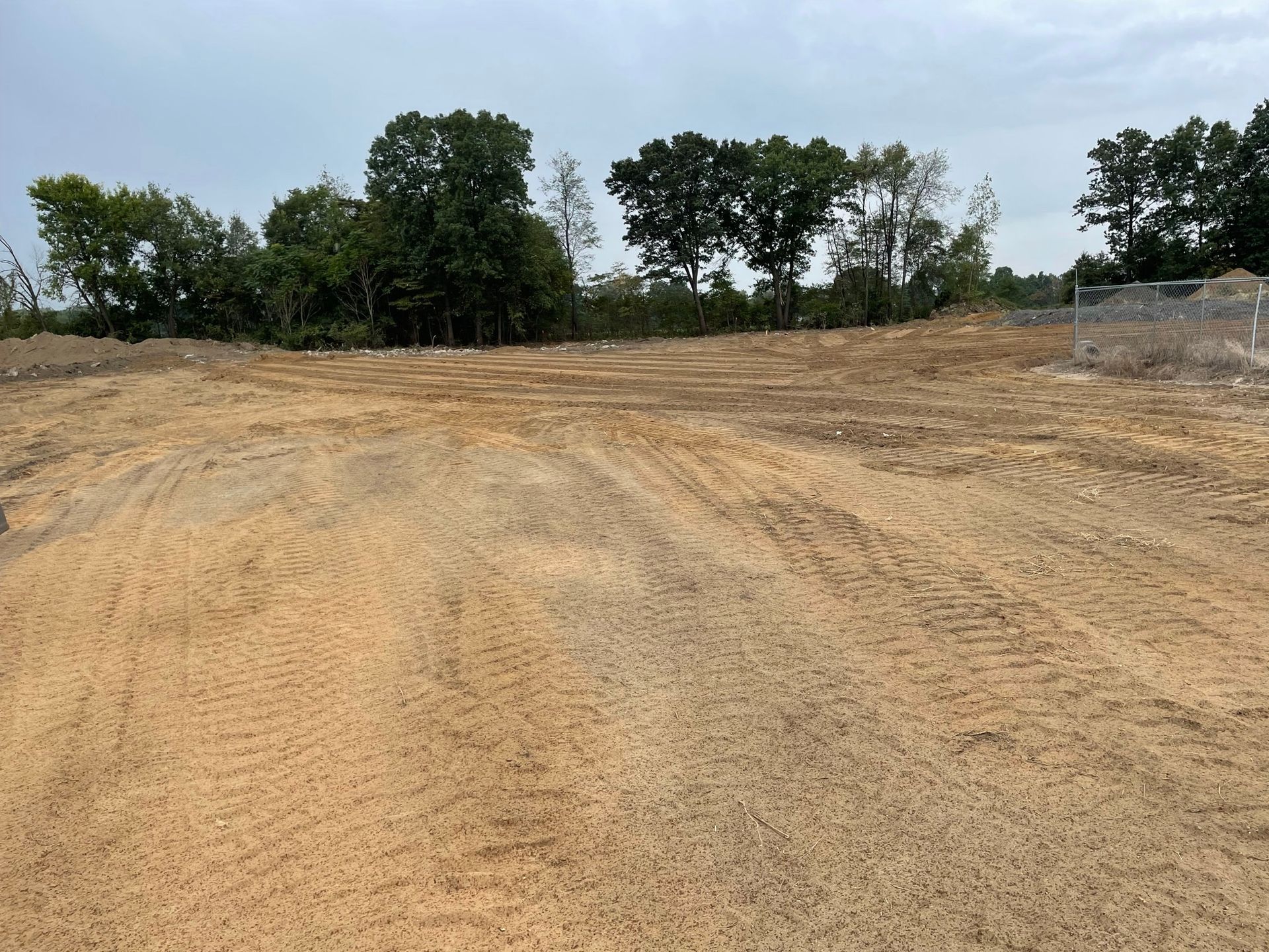 Cleared dirt lot with tire tracks in the foreground, trees and overcast sky in the background.