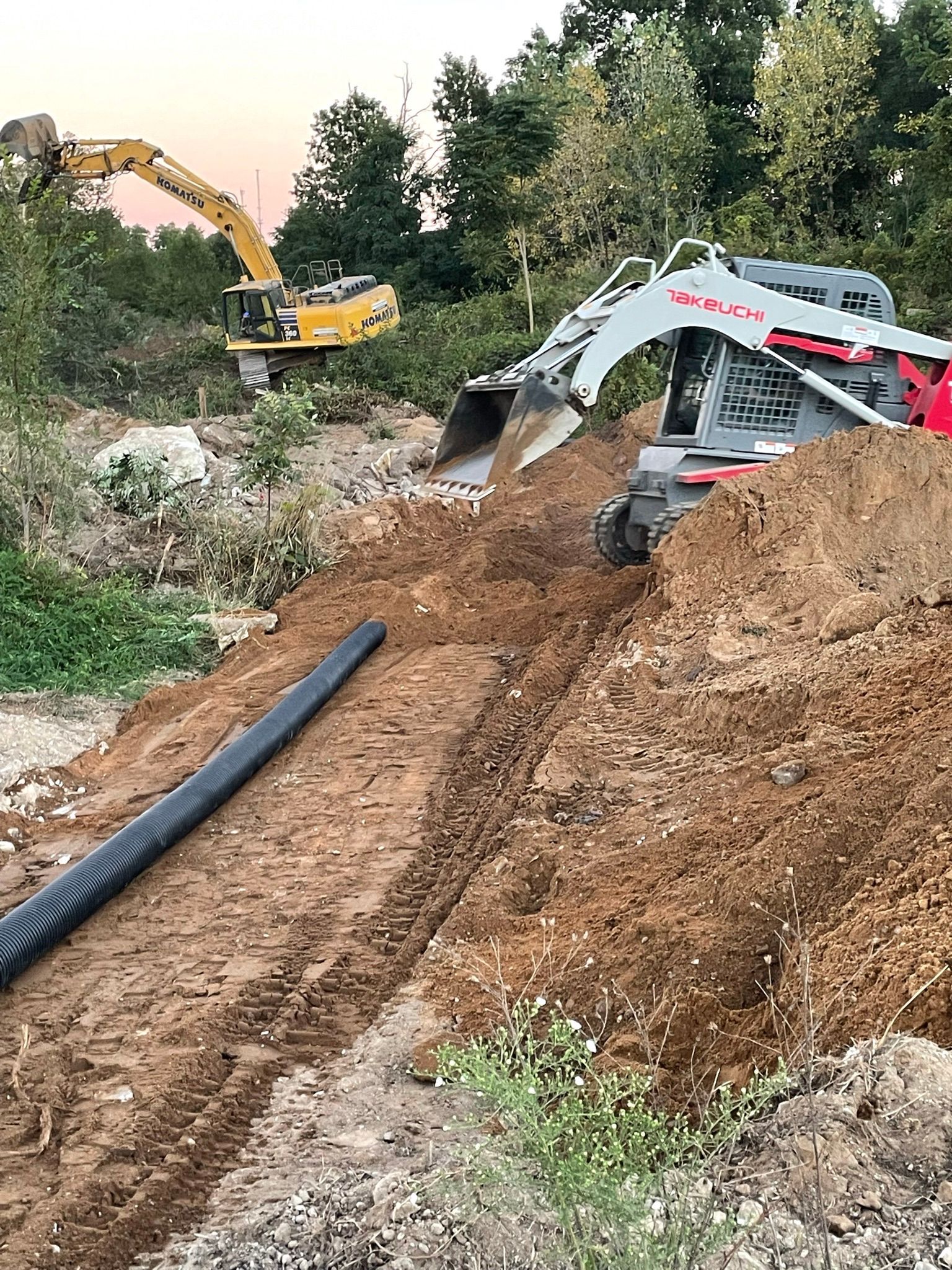Construction site with two excavators digging in dirt, a black pipe in a trench.