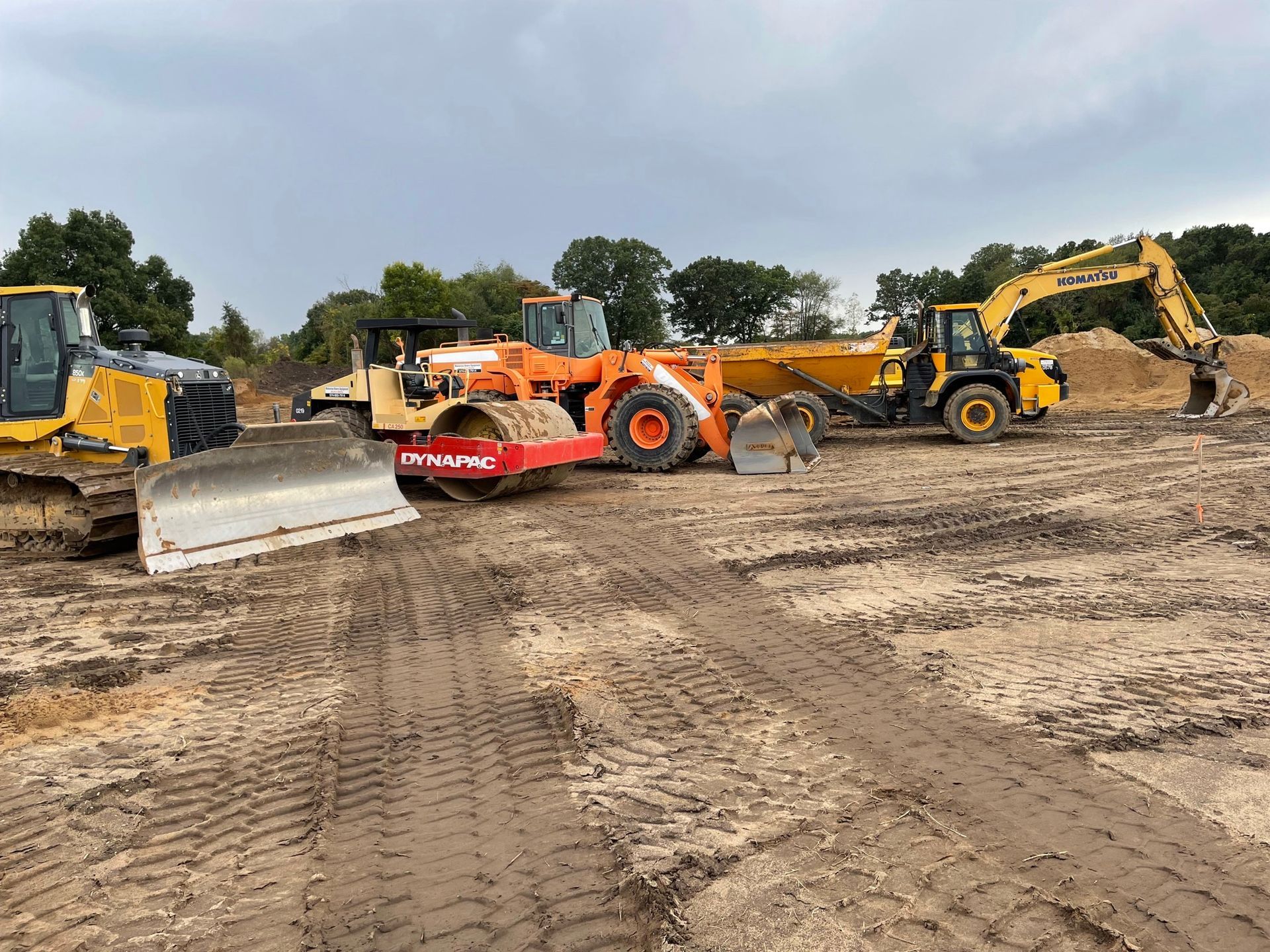 Construction equipment on a muddy construction site, including a bulldozer, roller, and excavator, under cloudy skies.