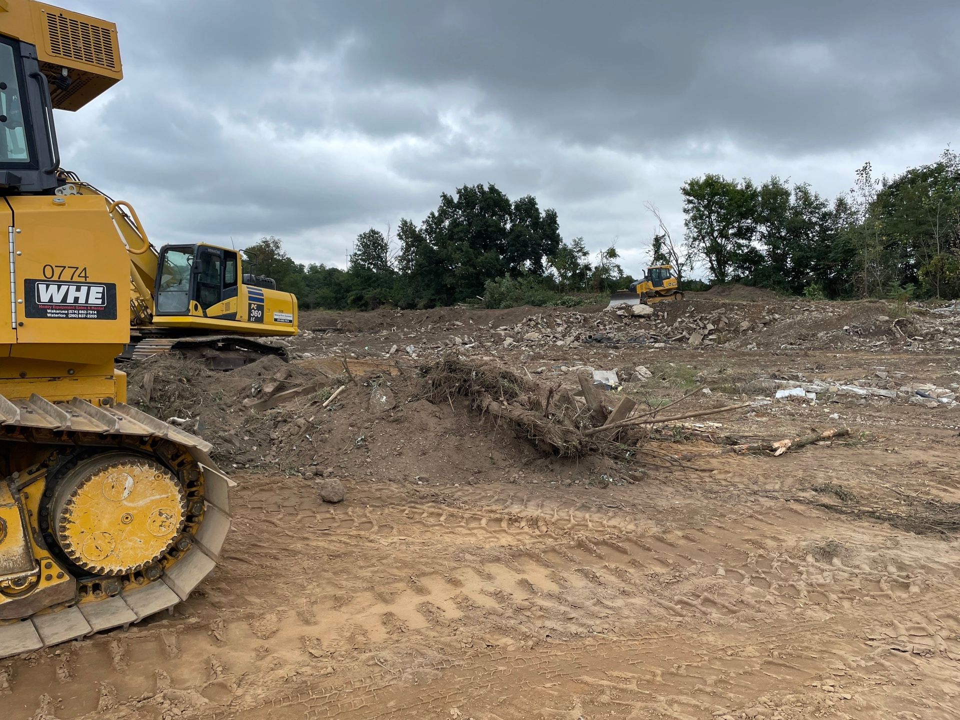 Yellow bulldozer and excavator on a construction site clearing land under a cloudy sky.
