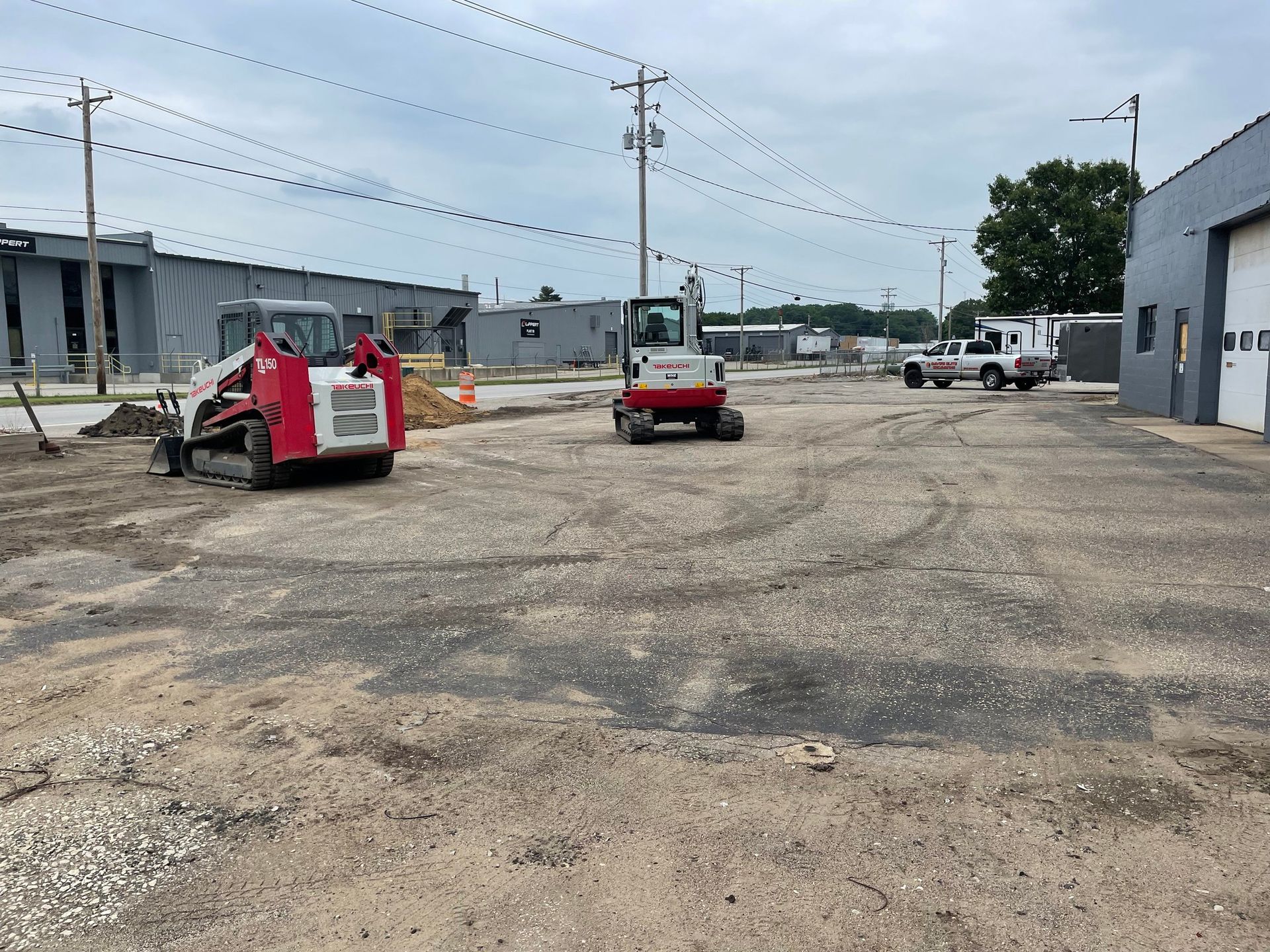 Construction site with two red and white Bobcat machines, and a truck parked nearby. Pavement is torn up.