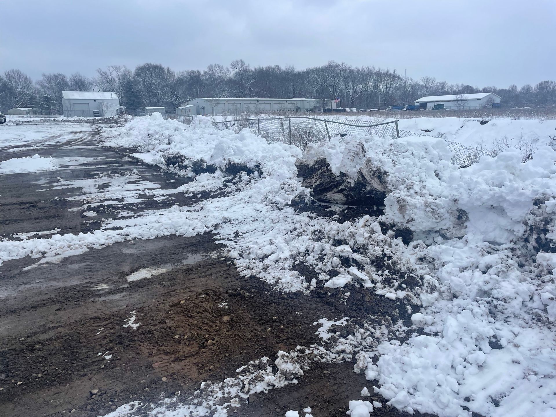 Snowy field with plowed dirt in the foreground, buildings and trees in the distance under a cloudy sky.
