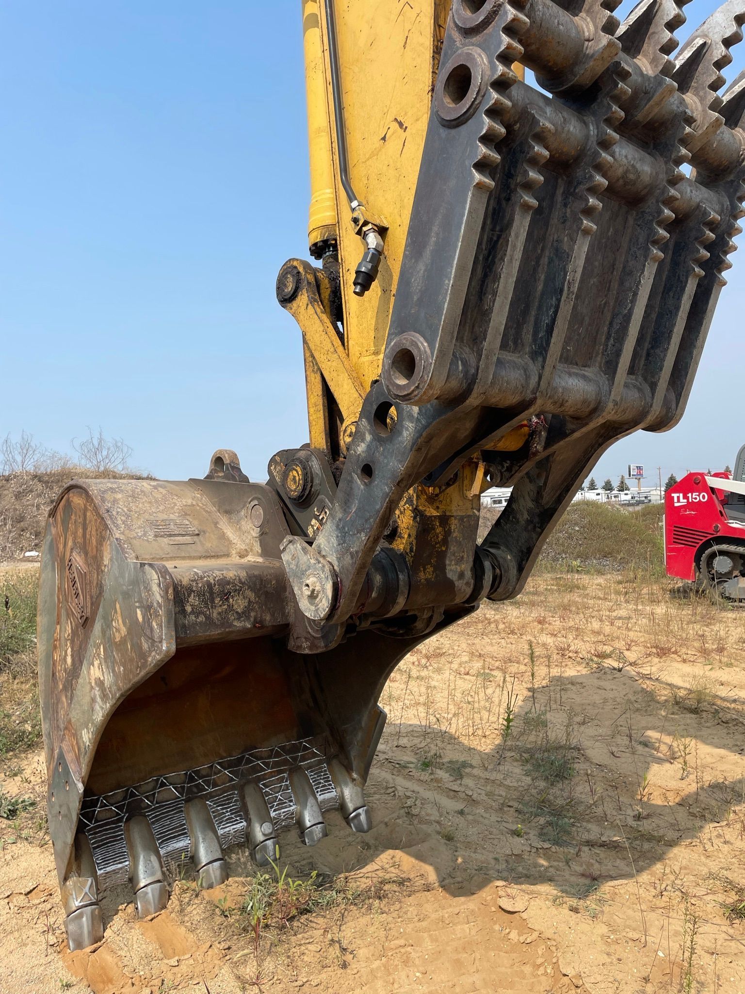 Yellow excavator bucket with teeth on dry, dirt ground. Bright sky in background.