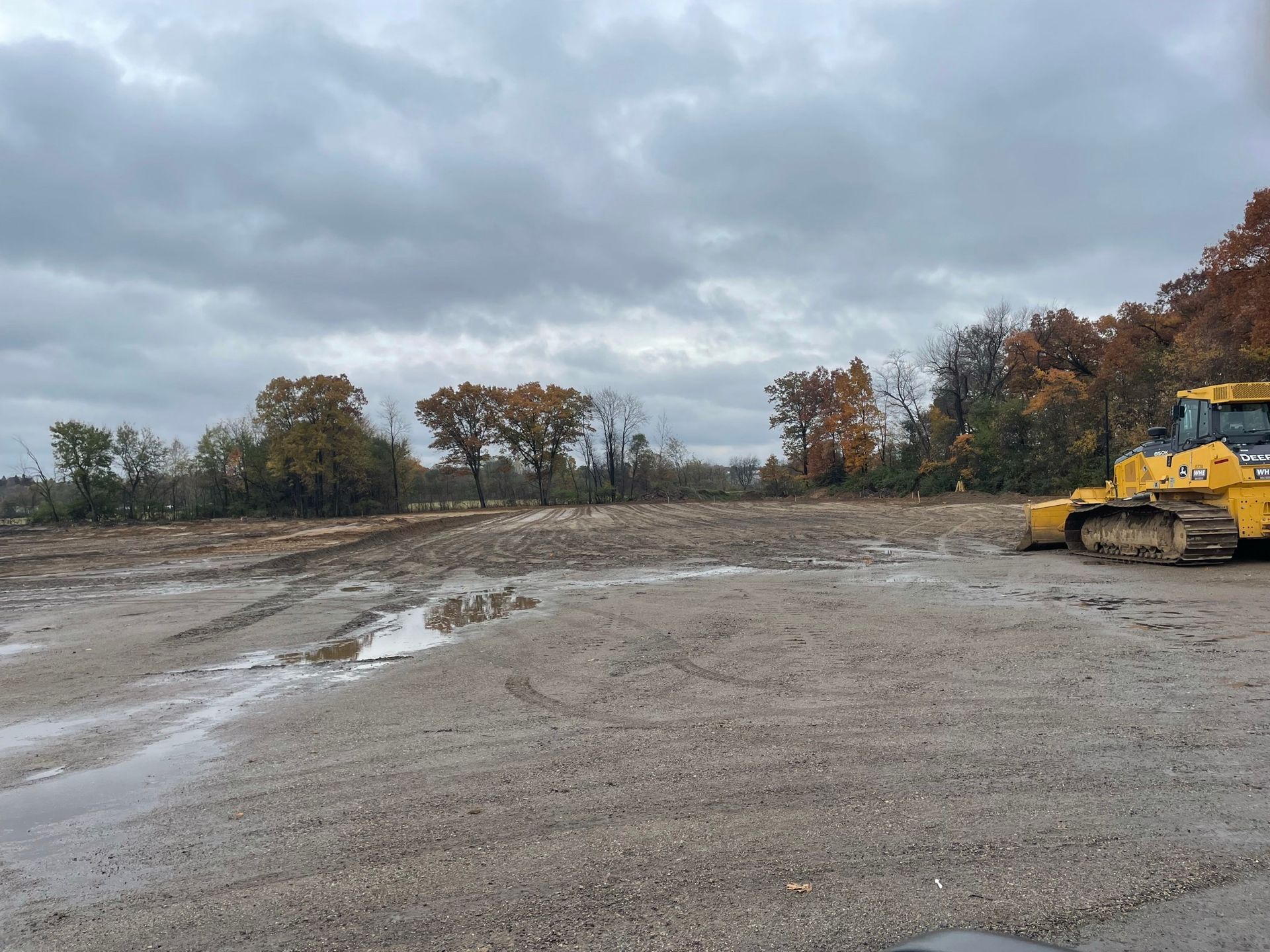 A construction site on an overcast day, with a bulldozer parked to the right. Bare ground and trees in the background.