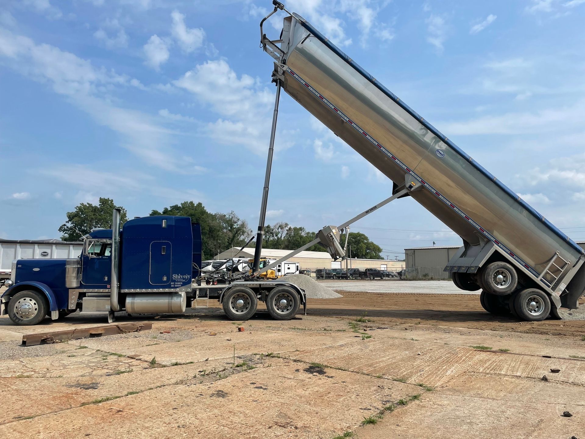 Blue semi-truck with tilted bed dumping gravel onto a dirt ground under a blue sky.