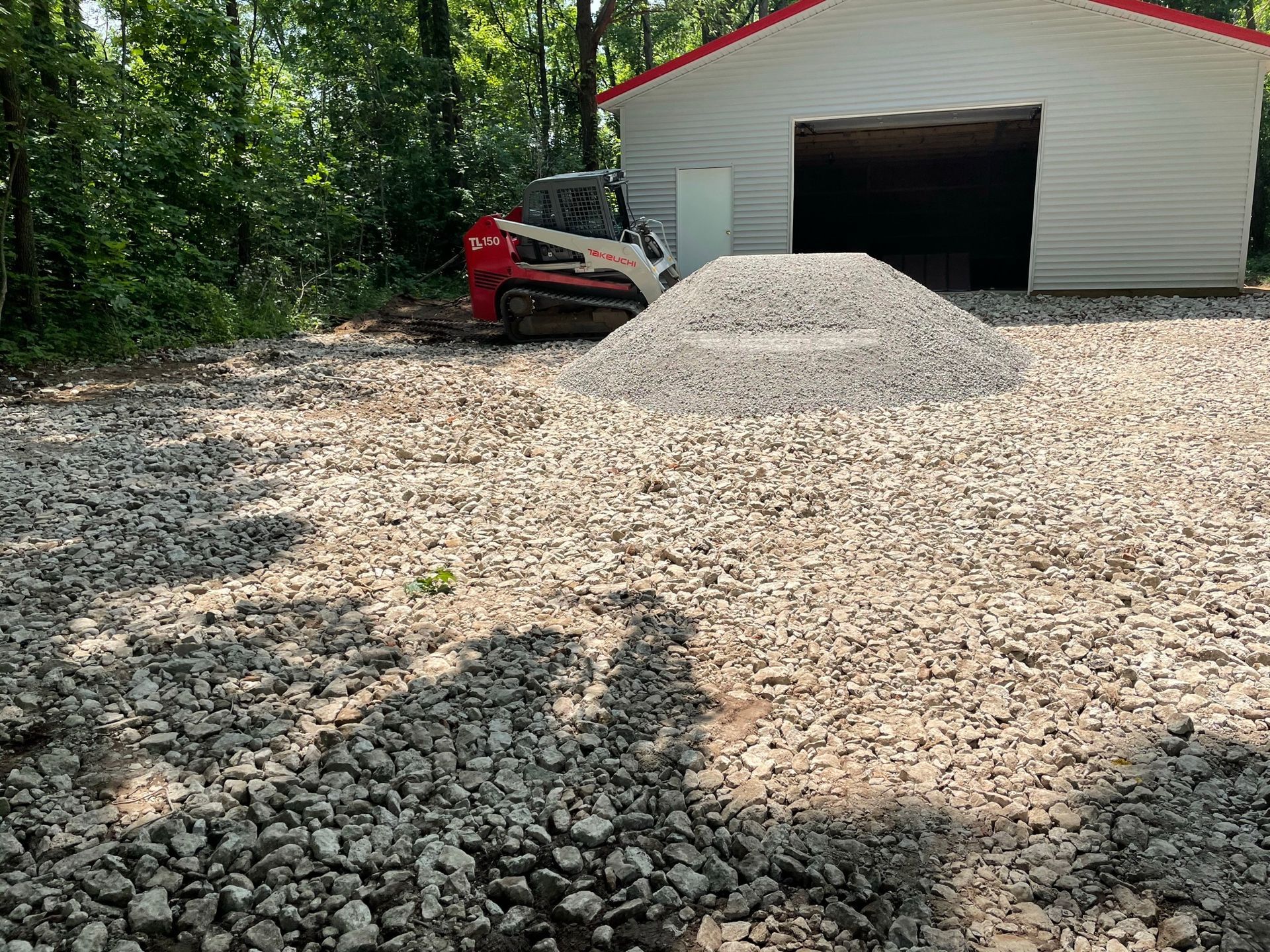 Gravel driveway with large pile of gravel; a red skid steer loader is near a white building with an open garage.