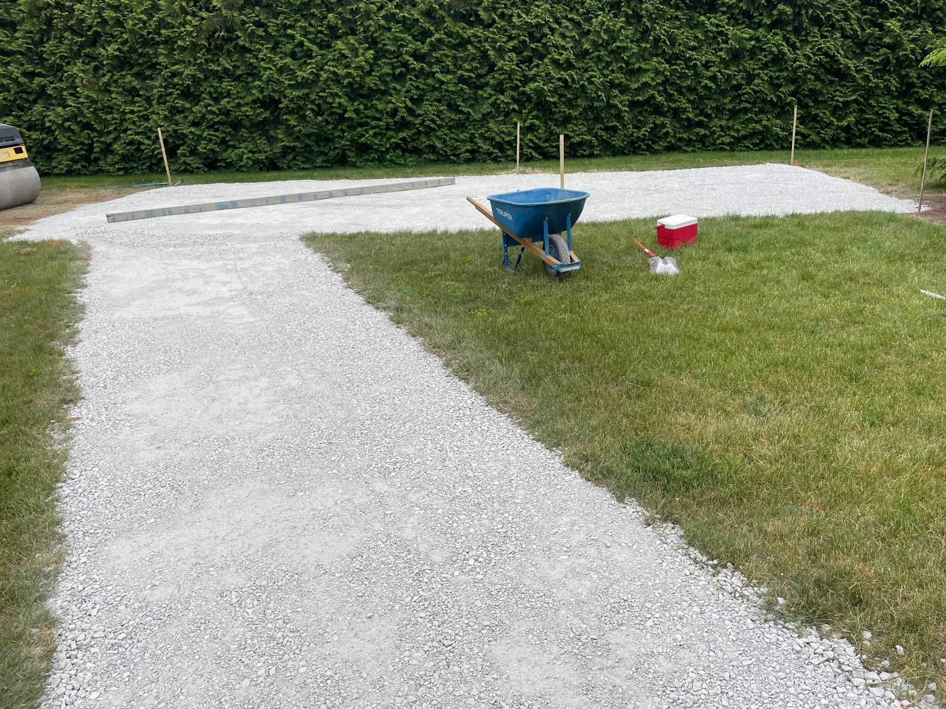 Gravel pathway and patio under construction on grass, with wheelbarrow and spreader in a backyard.