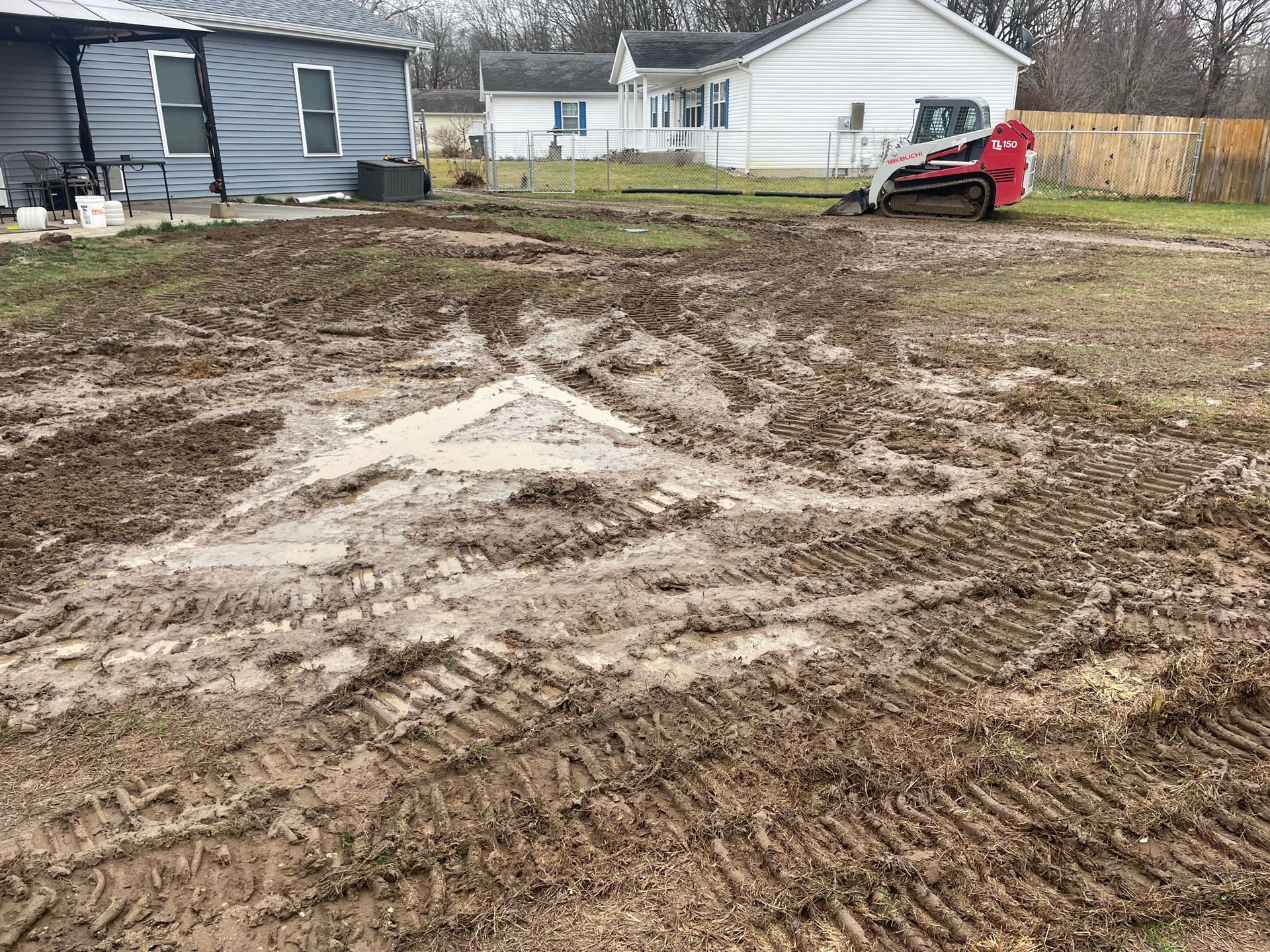 Muddy yard with tire tracks; a small tractor and two houses are visible in the background.