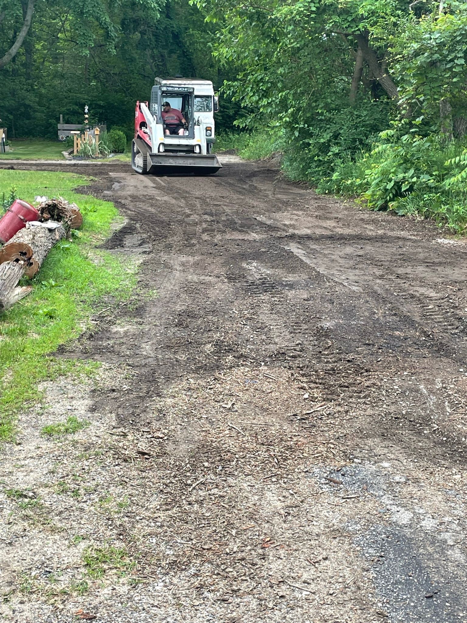 A small construction vehicle leveling gravel on a dirt driveway, beside a grassy area and trees.