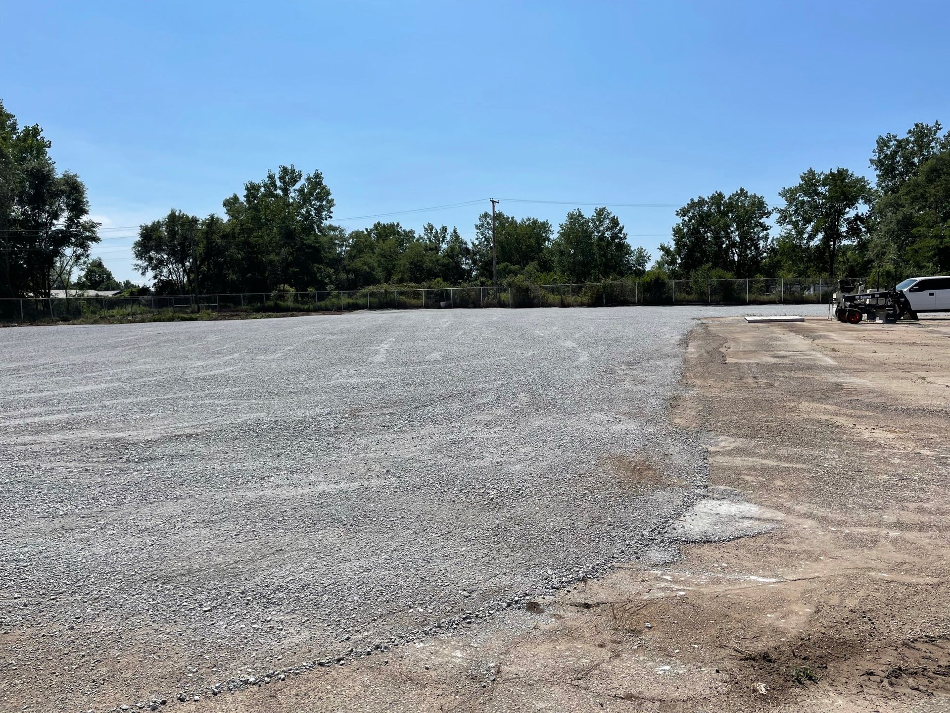 A gravel-covered lot under a bright blue sky, trees in the background, a small vehicle on the right.