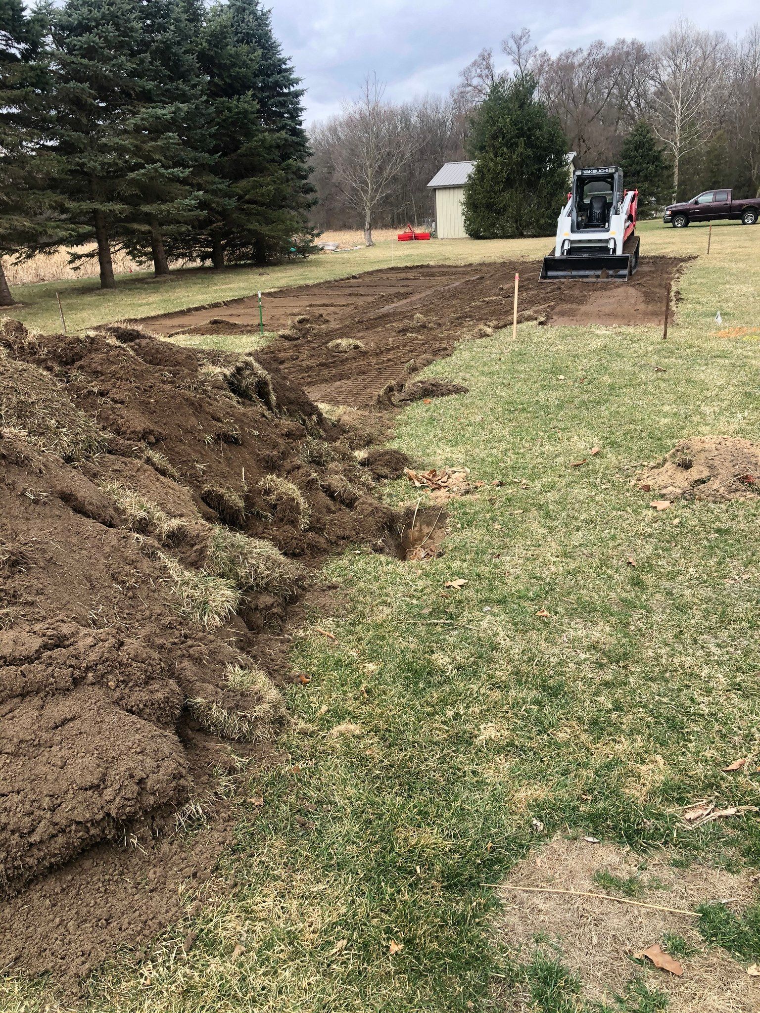 A small white excavator digs in a grassy field, with a long, dirt trench and pile of earth alongside.