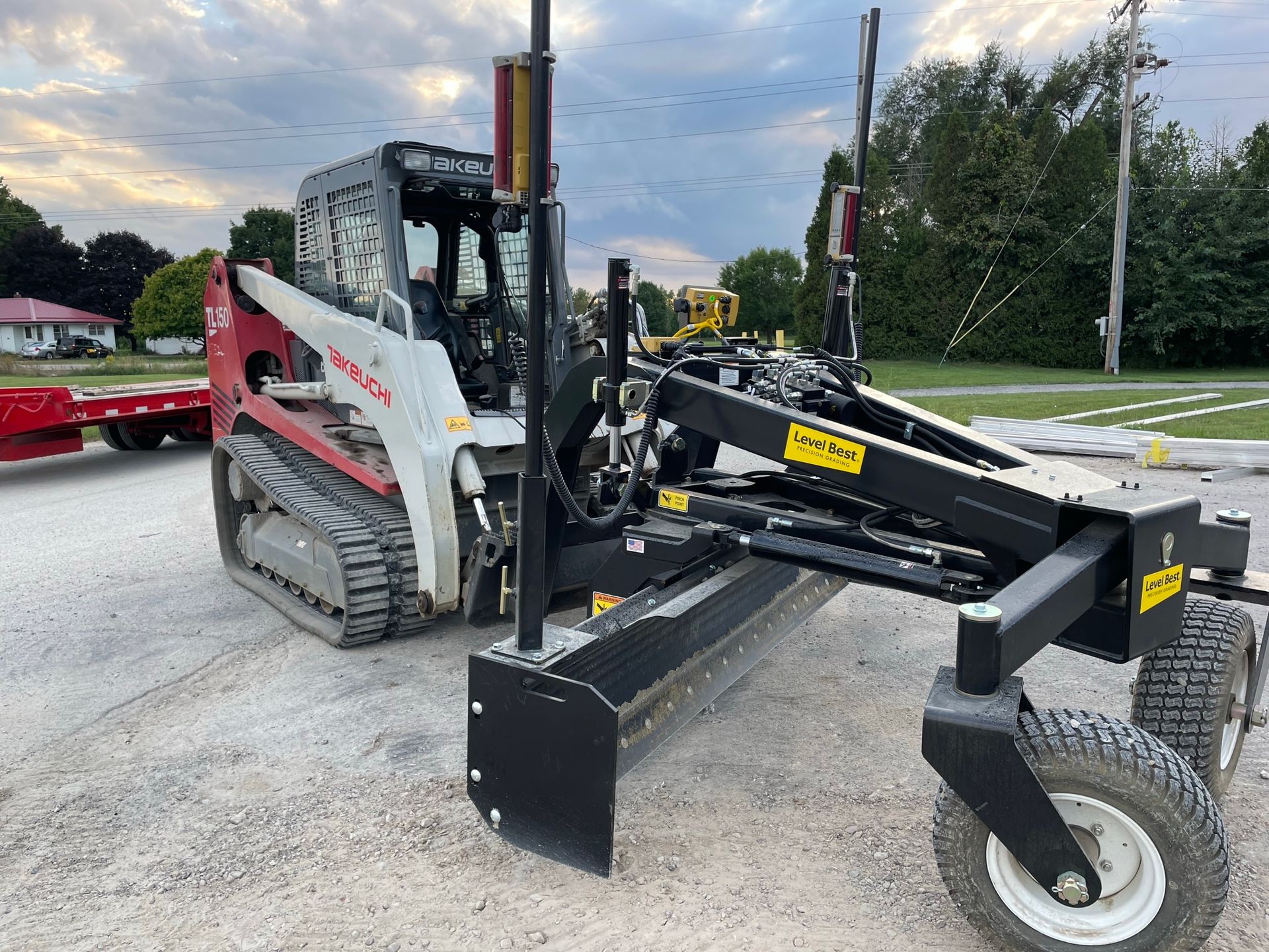 A skid steer loader with a grading attachment on a gravel surface outdoors.