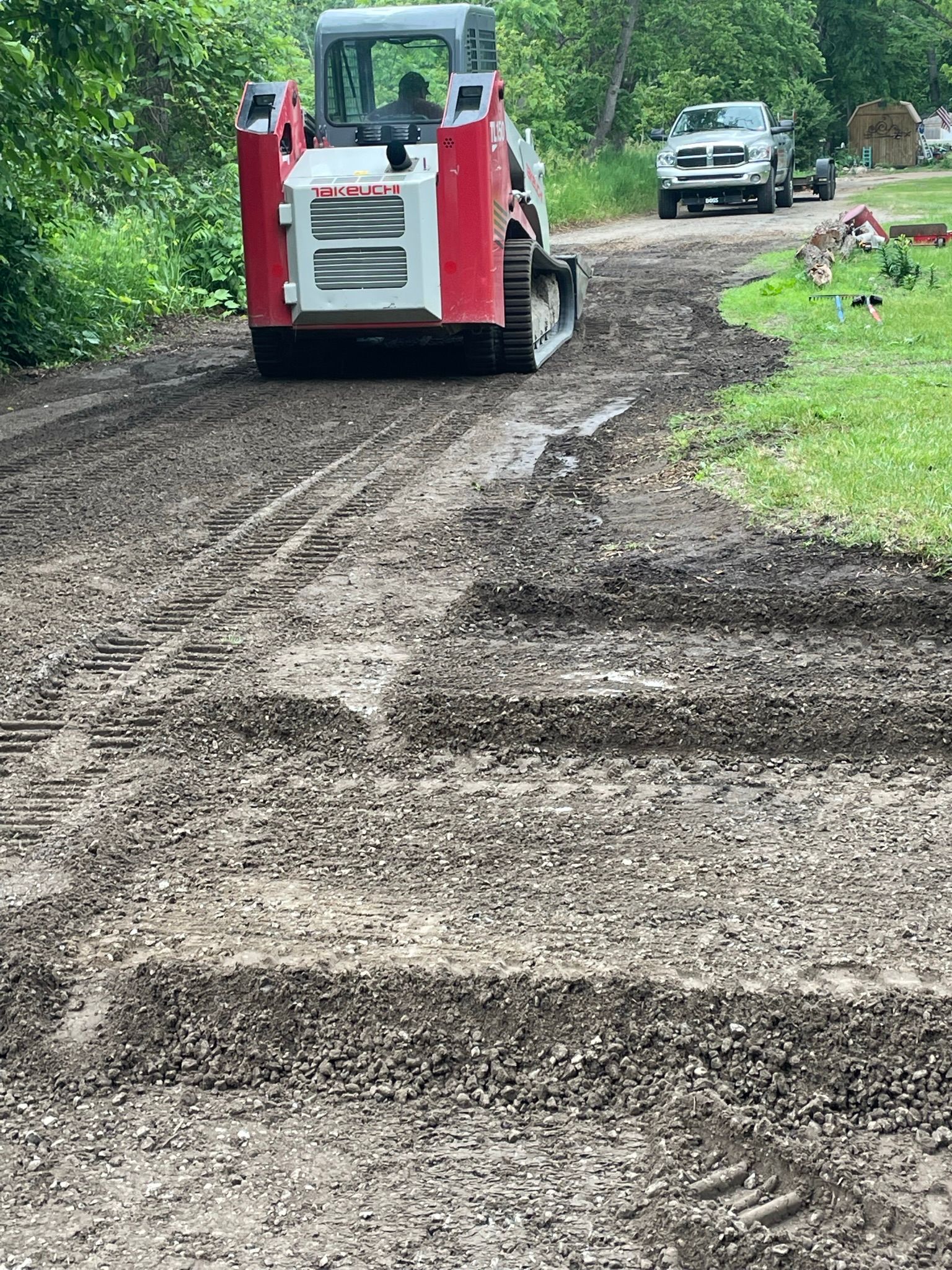 Skid steer grading gravel road. Red and white machine, light-colored pickup truck in background.